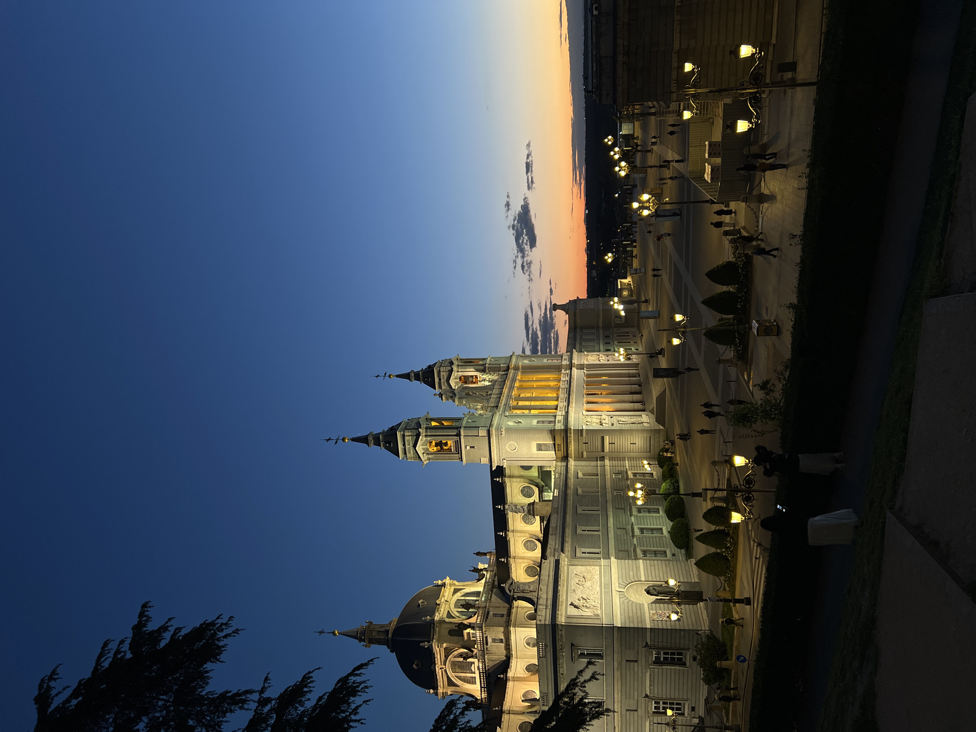 The image depicts the Almudena Cathedral in Madrid, Spain, at dusk. The sky transitions from deep blue to orange near the horizon. The cathedral is illuminated, showcasing its architectural details. A plaza with ornate lampposts and figures are visible in front of the cathedral. Pedestrians can be seen walking in the plaza. There are also people sitting along a walkway. The foreground includes dark trees.