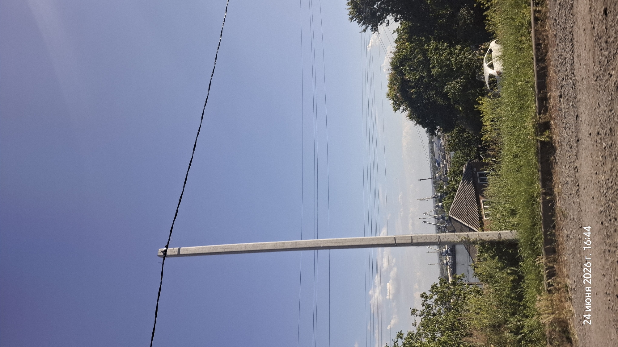 The media file is a vertically oriented photograph taken on a sunny day. The scene depicts an outdoor setting with a clear blue sky dominating the upper portion of the image. In the foreground, a tall, grey, rectangular concrete post or beam extends horizontally across the frame. Thin black wires, likely power lines, are visible stretching across the sky.
The mid-ground shows a lush green area with tall grass and trees. Partially visible amidst the greenery, on the left side, is a white car chassis. Further to the left, a body of water, possibly a river or bay, can be seen with several cranes and industrial structures in the distance, suggesting a port or industrial area. A small, dark-roofed building with a white-framed window is situated between the greenery and the water.
The right side of the image features a gravel road or path running diagonally into the distance. The overall impression is of a semi-rural or industrial landscape on a bright, clear day.
Visible text in the lower right corner reads "24 ИЮНЯ 2026 г. 16:44", indicating the date and time the photograph was taken in Russian: June 24, 2026, 4:44 PM. The location context provided is Rostov-on-Don, Russia.