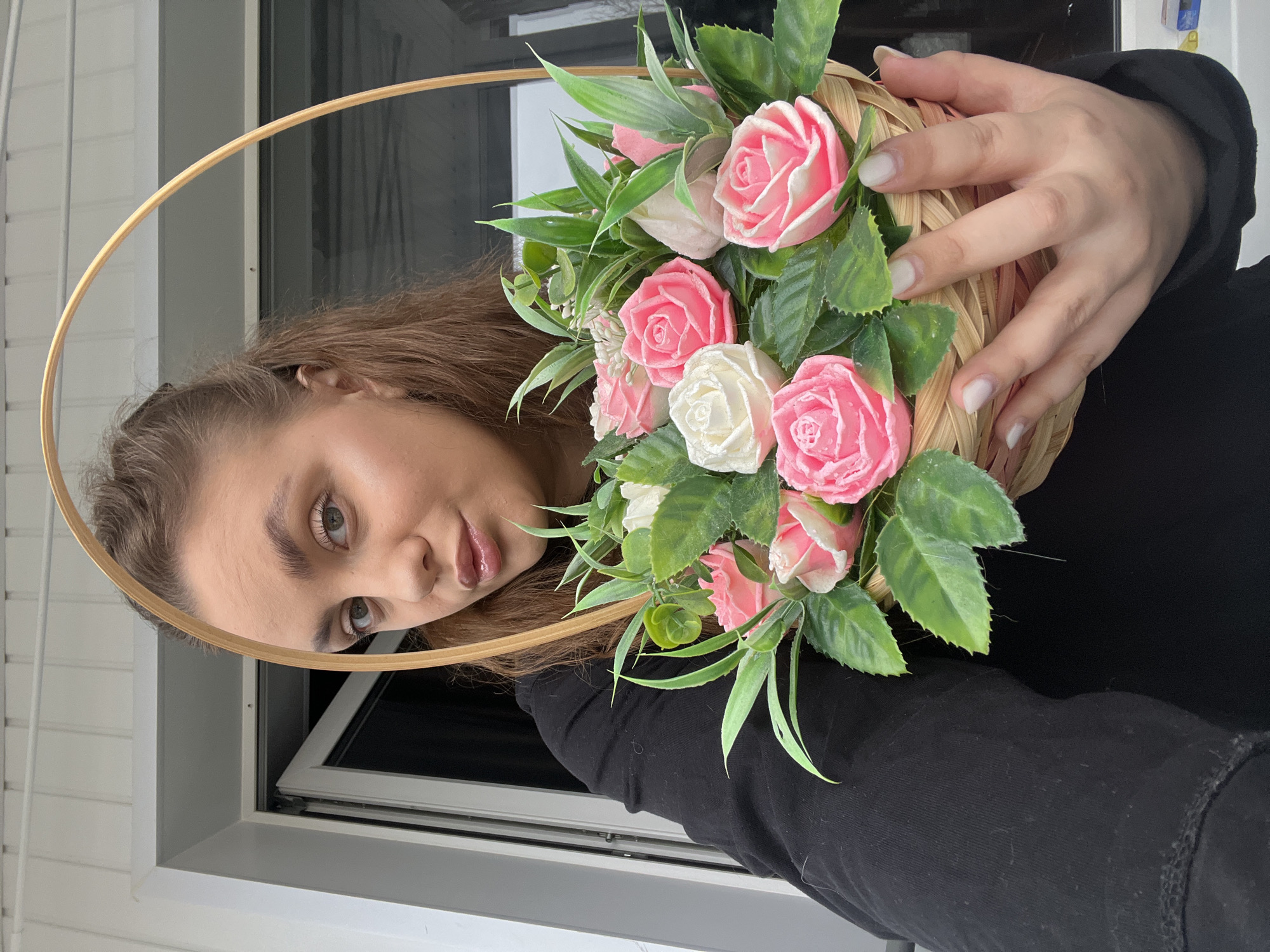 A woman is holding a basket of flowers. The basket is woven and contains pink and white roses with green leaves. The woman is wearing a black shirt. She has brown hair and light eyes. She is positioned in front of a window. The location is identified as Chaikovsky, Russia, based on the context. The scene appears to be indoors.