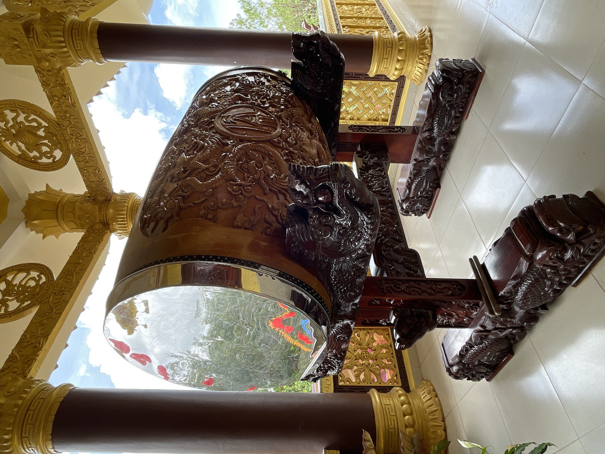 A large, intricately carved wooden drum rests on a dark red wooden stand, also adorned with dragon carvings. The drum has a clear, reflective surface on one end, through which trees and sky are visible. Inside the drum, red and orange decorative elements, possibly paper cutouts of dragons, are visible. The drum and stand are positioned on a tiled floor under a covered walkway supported by large, dark brown pillars with gold accents. Gold decorative elements are also visible on the ceiling and railings. The scene appears to be outdoors, with a bright sky and some greenery visible in the background. The location is identified as the Linh Phuoc Pagoda in Da Lat, Vietnam.