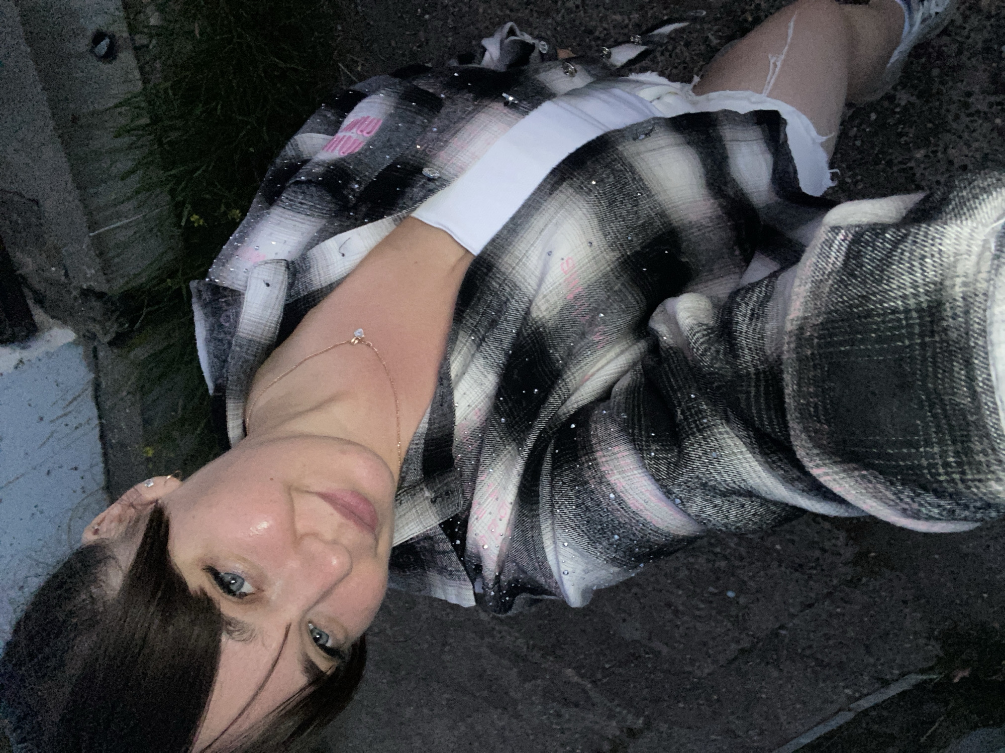 A woman is taking a selfie outdoors at night in Votkinsk, Russia. She is wearing a plaid shirt over a white top and ripped denim shorts. The ground is paved and there is grass and a concrete structure in the background.