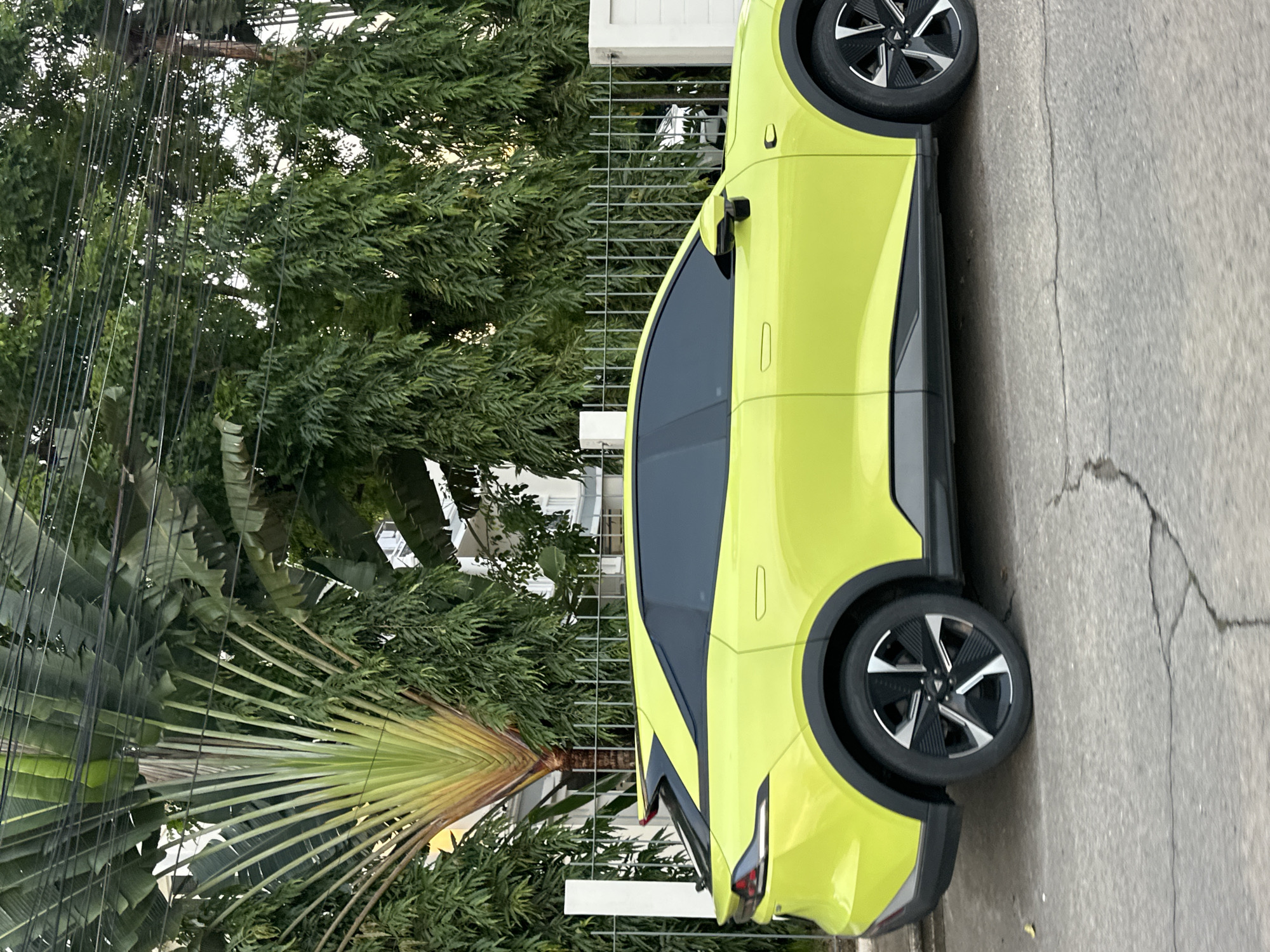 An eye-level, medium shot captures the side profile of a bright yellow electric vehicle parked on a cracked asphalt road in Kathu, Thailand. The car, a modern SUV, has black tinted windows, black wheel arches, and a distinctive two-tone wheel design with silver and black spokes. Behind the car, a silver metal fence separates the property from the street. Beyond the fence, lush green foliage of trees and a large fan palm dominate the background. The building in the background is a white house with multiple windows, partially obscured by the vegetation. Overhead, a network of black electrical wires crisscrosses the frame, further emphasizing an urban or semi-urban setting. The lighting suggests it is daytime, with soft light illuminating the scene. There are no people visible in the image.