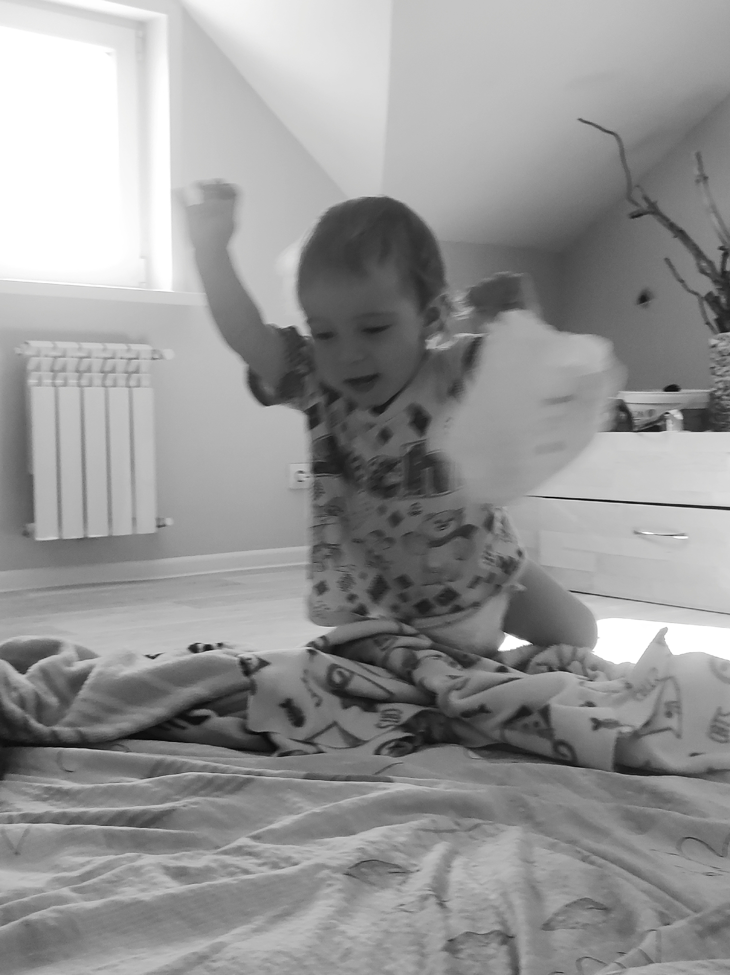 A black and white image shows a toddler in a patterned t-shirt and diaper kneeling on a bed covered with patterned blankets. The child has one arm raised in the air with a blurred object in hand. Behind the child, there is a white radiator and a window on the left. A white dresser with a vase and dried branches on top is visible on the right. The environment appears to be an indoor room, likely a bedroom. There is no mention of Фоки, Russia.