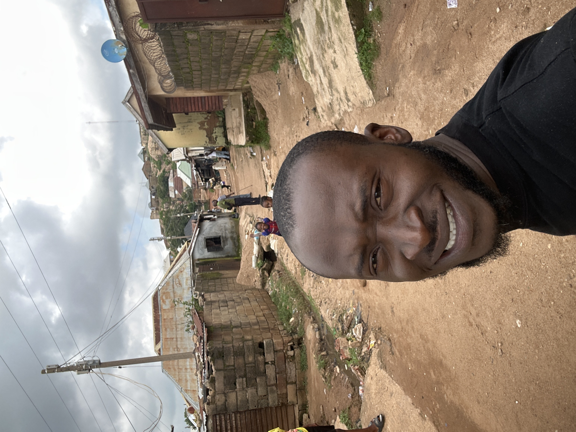 A man with dark skin and a beard takes a selfie in the foreground, smiling and looking towards the camera. He is wearing a black t-shirt. In the background, a dirt road winds through a residential area. Several structures, some with corrugated metal roofs and others made of brick, line the road. A blue satellite dish is visible on top of one of the buildings. A few other people, including two children, are present in the middle ground, walking along the road. Electrical poles and wires are prominent overhead. The sky is overcast with clouds. The scene appears to be in a somewhat informal settlement, typical of parts of Jos, Nigeria.