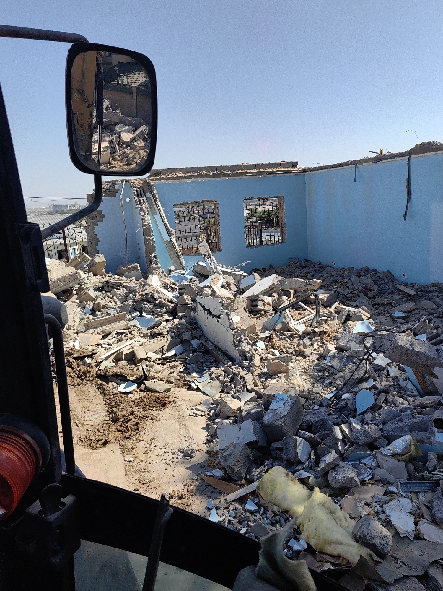 The image captures a scene of demolition in Shymkent, Kazakhstan. The perspective is from within a construction vehicle, looking out at a partially destroyed building. The building's interior is exposed, revealing blue walls and piles of debris, including concrete, bricks, and insulation. The exterior is visible through a side mirror, reflecting more rubble and a clear blue sky. The overall environment suggests an active demolition site.