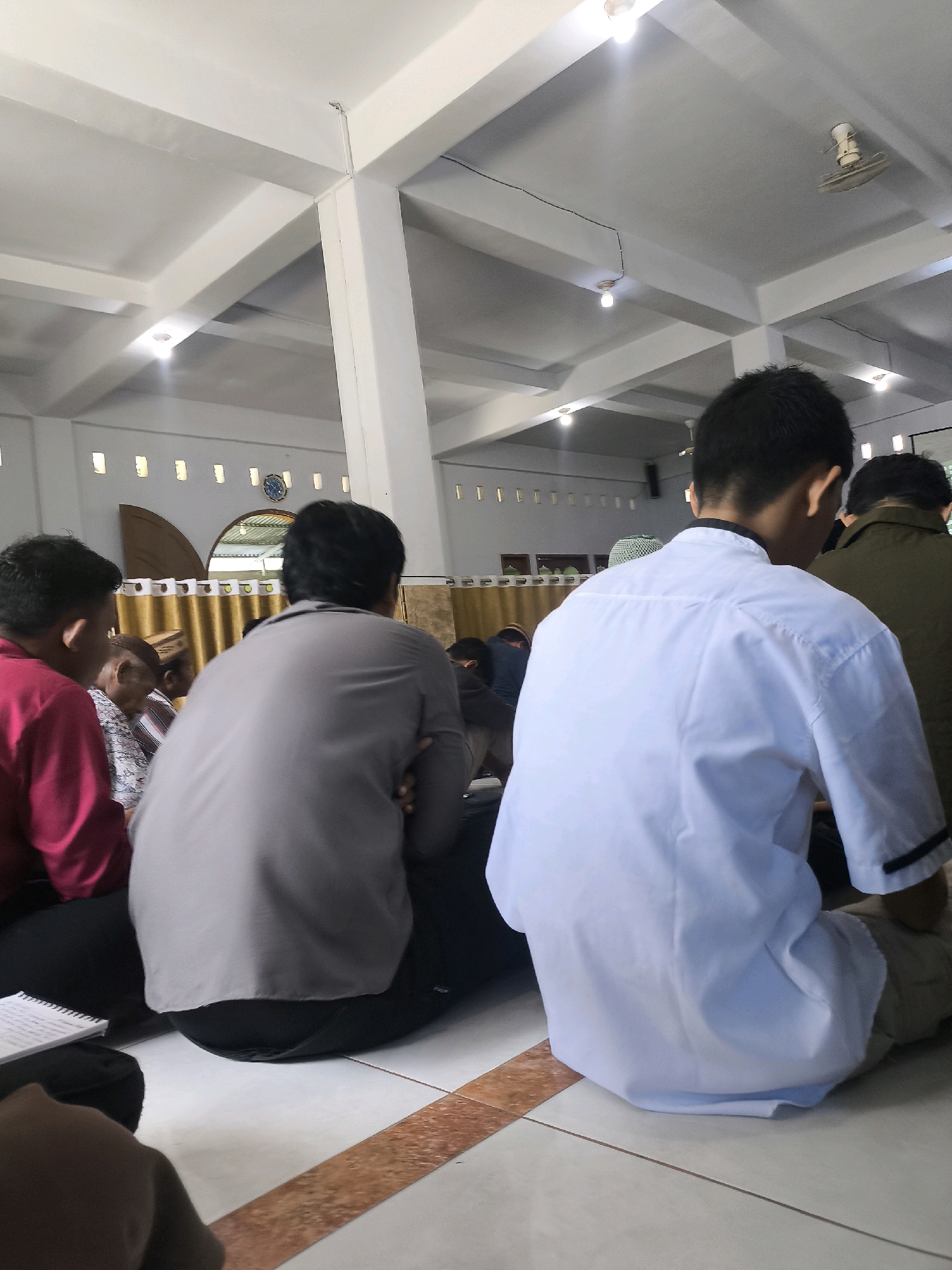The image depicts the interior of what appears to be a mosque or prayer hall in Gorontalo, Indonesia. Several individuals are seen sitting on the floor, facing away from the camera. One person in the foreground is wearing a white long-sleeved shirt and light-colored pants. To their left, another person is wearing a grey shirt and dark pants. Further to the left, a person in a red shirt is visible. The ceiling features a white, angled beam structure with lights. Along the back wall, a decorative arched window is present above a patterned curtain. The floor is tiled with a lighter color and accented by a brown stripe. The overall atmosphere suggests a religious gathering or prayer session.