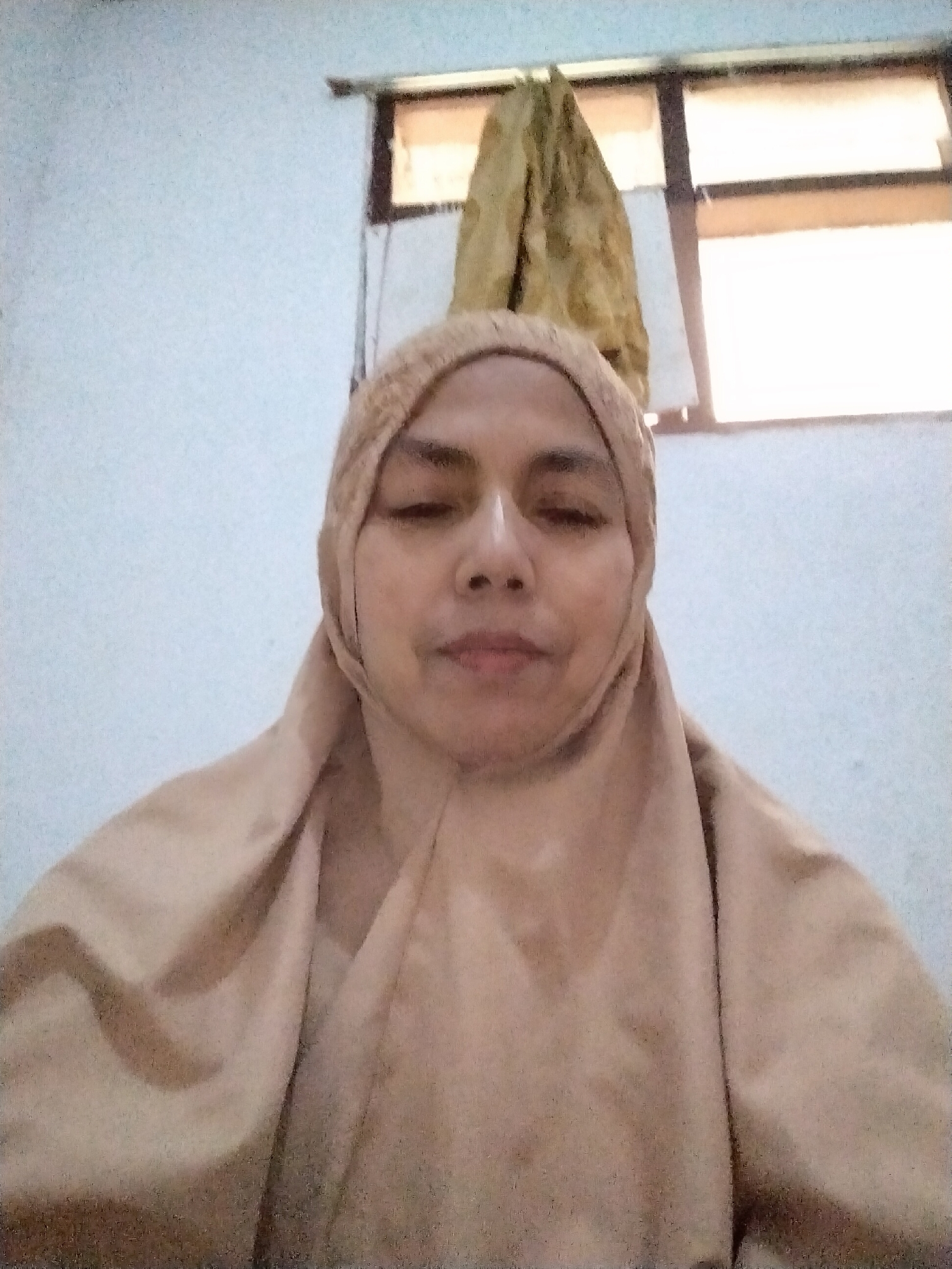 A frontal, medium shot shows a woman wearing a light brown hijab and a matching garment that covers her head and body. Her eyes are closed and her mouth is slightly open. She is positioned centrally in the frame against a plain, off-white wall. In the background, a window is visible, divided into two panes by a horizontal bar. The upper pane is partially obscured by a draped, yellowish-brown fabric that hangs from above, possibly a curtain or a piece of clothing. The lighting in the scene appears to be coming from the window, casting a soft glow. The image was taken indoors, likely in a residential setting. The location specified is Sukamanah, Indonesia, though this is not visually evident from the image itself.