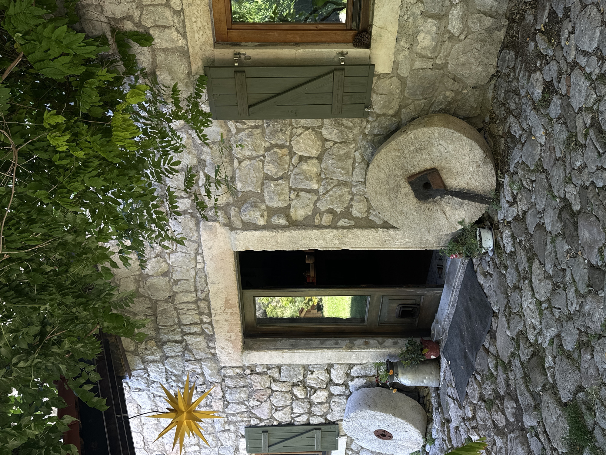An exterior view in Bartula, Montenegro, depicts the stone facade of a building partially obscured by dense green foliage overhead. The building features walls constructed from irregular, light-colored stone blocks. A central wooden door, painted greenish-brown with a glass panel, is ajar, reflecting green foliage and a distant structure; a dark interior is visible. The doorway is framed by large, worn stone blocks.
To the left of the door, a closed dark green wooden shutter covers a window opening. To the right, another dark green wooden shutter is slightly ajar, revealing a wooden window frame and a glass pane reflecting greenery. A small pinecone rests on the sill below this right window. Suspended from above, to the left of the doorway, is a large, multi-pointed yellow star decoration.
In front of the building, the ground is paved with irregular grey-brown cobblestones, with sparse weeds growing between them. A rectangular, dark grey doormat is positioned directly in front of the open door.
Two large, round, light-grey stone millstones lean against the building's facade. The millstone on the left has a reddish-brown metal hub; the millstone on the right features a dark, L-shaped metal fitting. Several plants in pots and vessels decorate the area: a tall, grey metal milk churn contains plants with small orange flowers near the left millstone, a smaller reddish-brown pot with green plants is next to it, and a white pot with a small plant and a pink flower is to the right of the doormat.
The scene is lit by natural daylight, with significant shadows cast by the overhead foliage, creating a dappled effect. No people are visible.