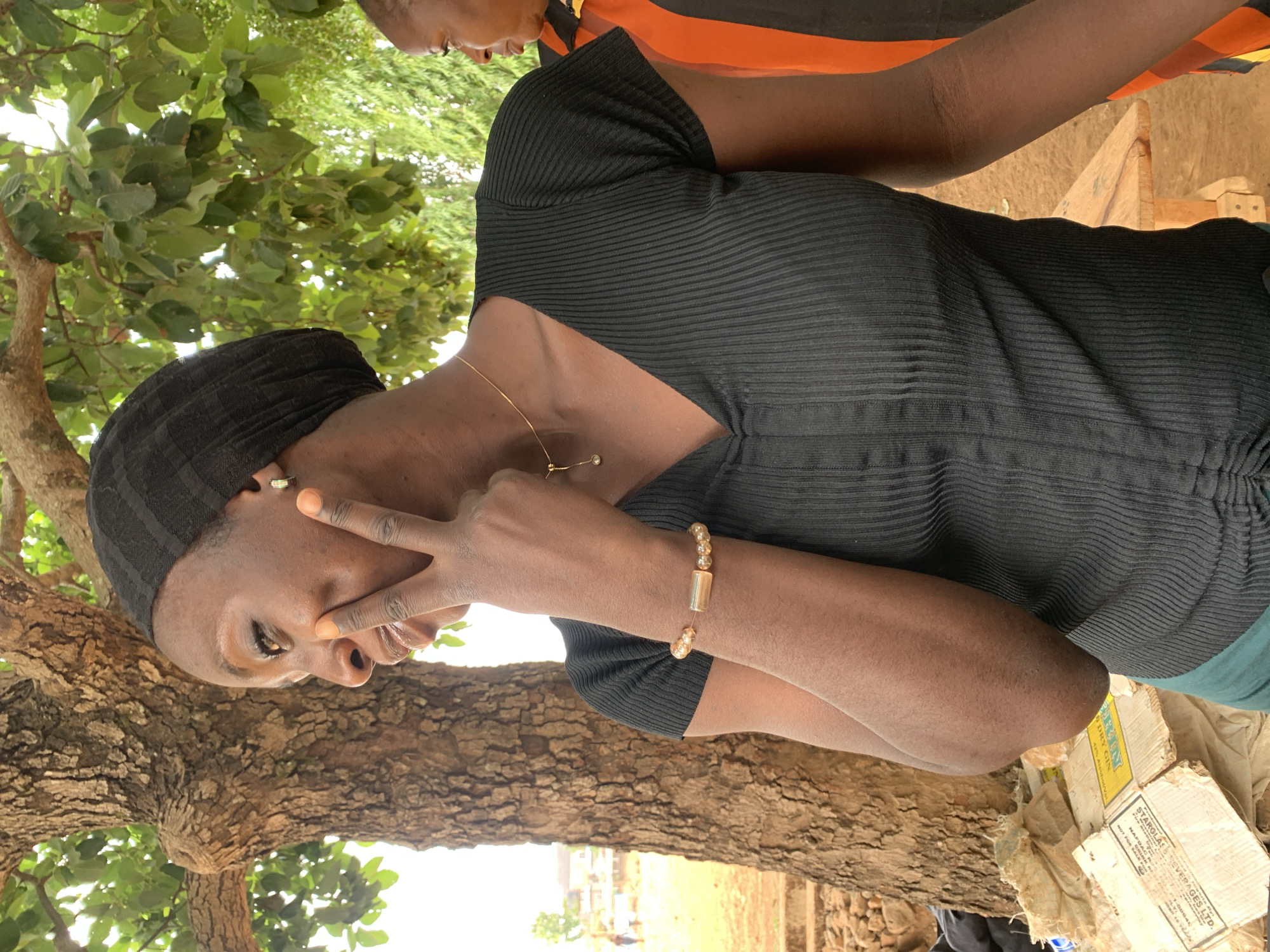 The image captures a woman outside, likely in Maigizo, Nigeria, based on the product details on the discarded packaging. She is wearing a black ribbed top, a black head covering, a gold necklace, and a beaded bracelet. The woman is holding up two fingers in a peace sign, partially obscuring her face.
The environment includes a large tree with visible bark texture and green foliage, suggesting an outdoor setting. There's also what appears to be a discarded cardboard box and other discarded items. Another person is partially visible in the background. The background is a field with a few scattered structures.