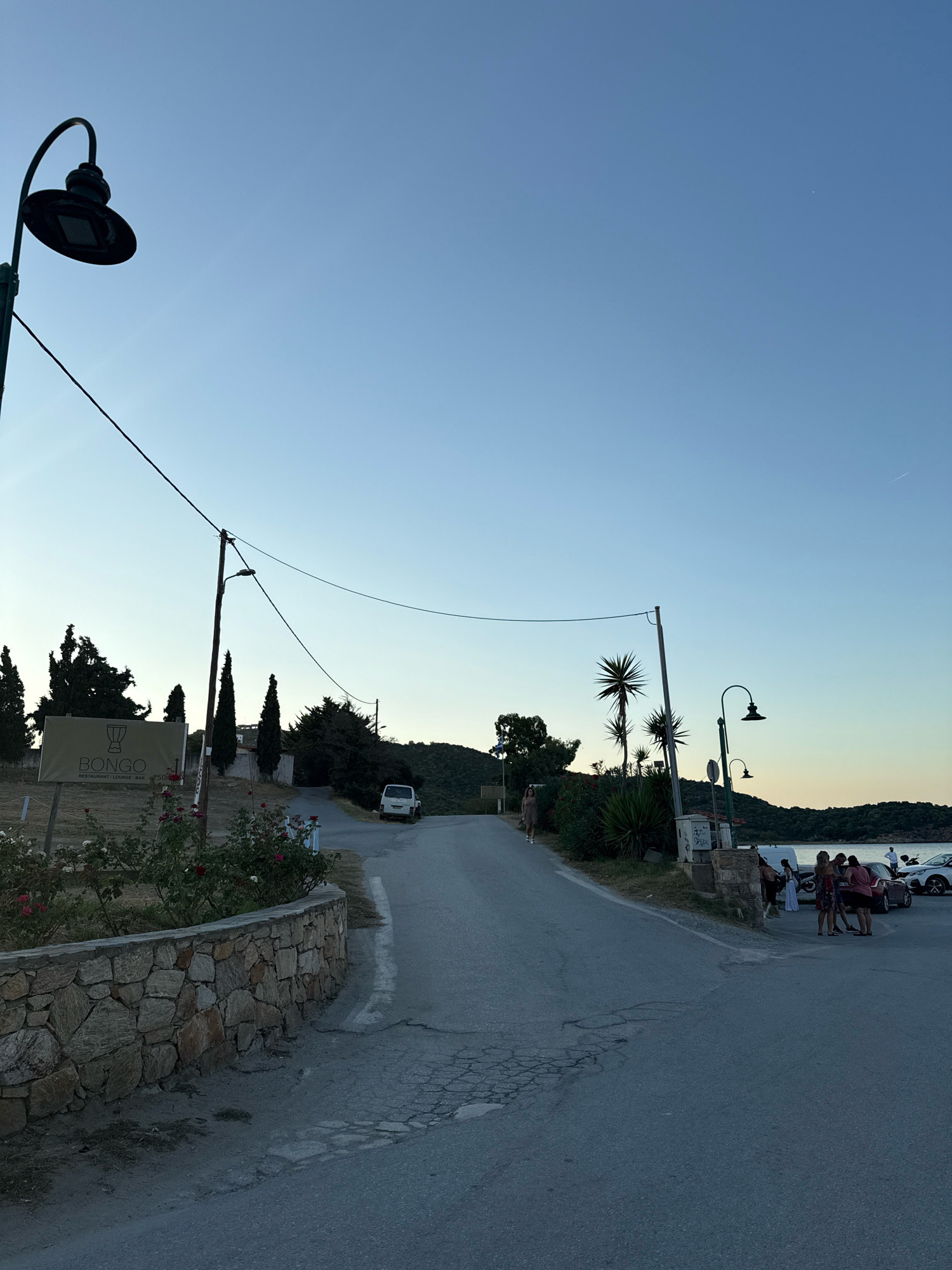 The image depicts a street scene in Ammouliani, Greece, at dusk. A paved road curves uphill, bordered by a low stone wall and lush vegetation on the left, and a gentle slope with trees and a sign for "BONGO RESTAURANT LOUNGE BAR" on the right. A tall, dark lamppost with an ornate fixture stands on the left, and another similar lamppost is visible further up the road on the right. Power lines traverse the sky.
On the right side of the road, near where it widens into what appears to be a parking area, a group of approximately six to eight individuals are gathered, some standing around vehicles. One person is seen walking uphill on the left side of the road. A white van is parked further up the road on the right. The sky is clear and a pale blue, transitioning to a warmer hue near the horizon, indicating the setting sun. The background features a body of water and a tree-covered hillside.