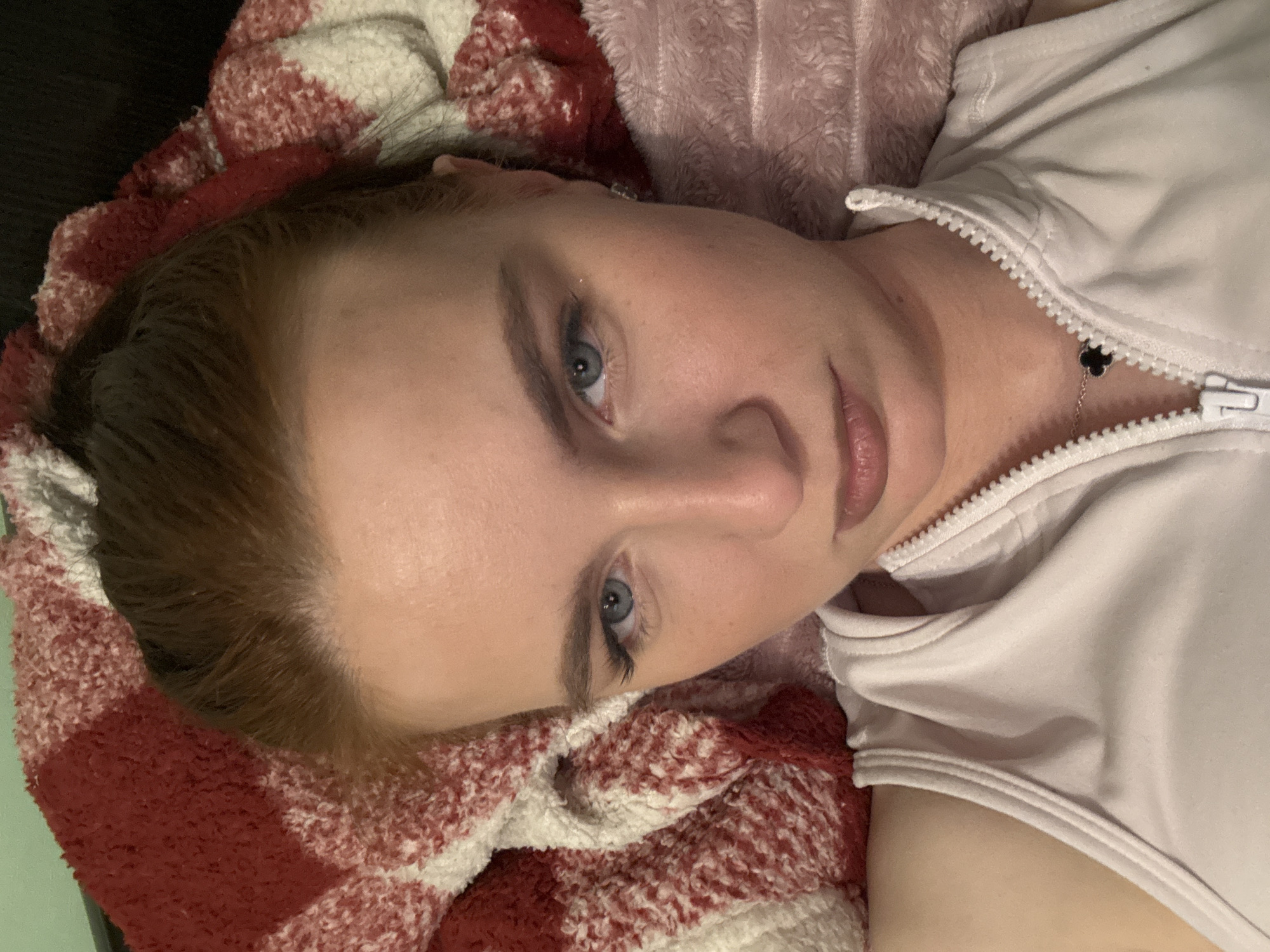A woman with light skin and blue eyes is lying on a bed covered with blankets of red and white plaid, and light pink. The woman is wearing a light pink zipper-front top and a thin necklace with a dark clover pendant. Her hair is light brown. The environment is indoors. There is no visible indication of Sinyavino, Russia.