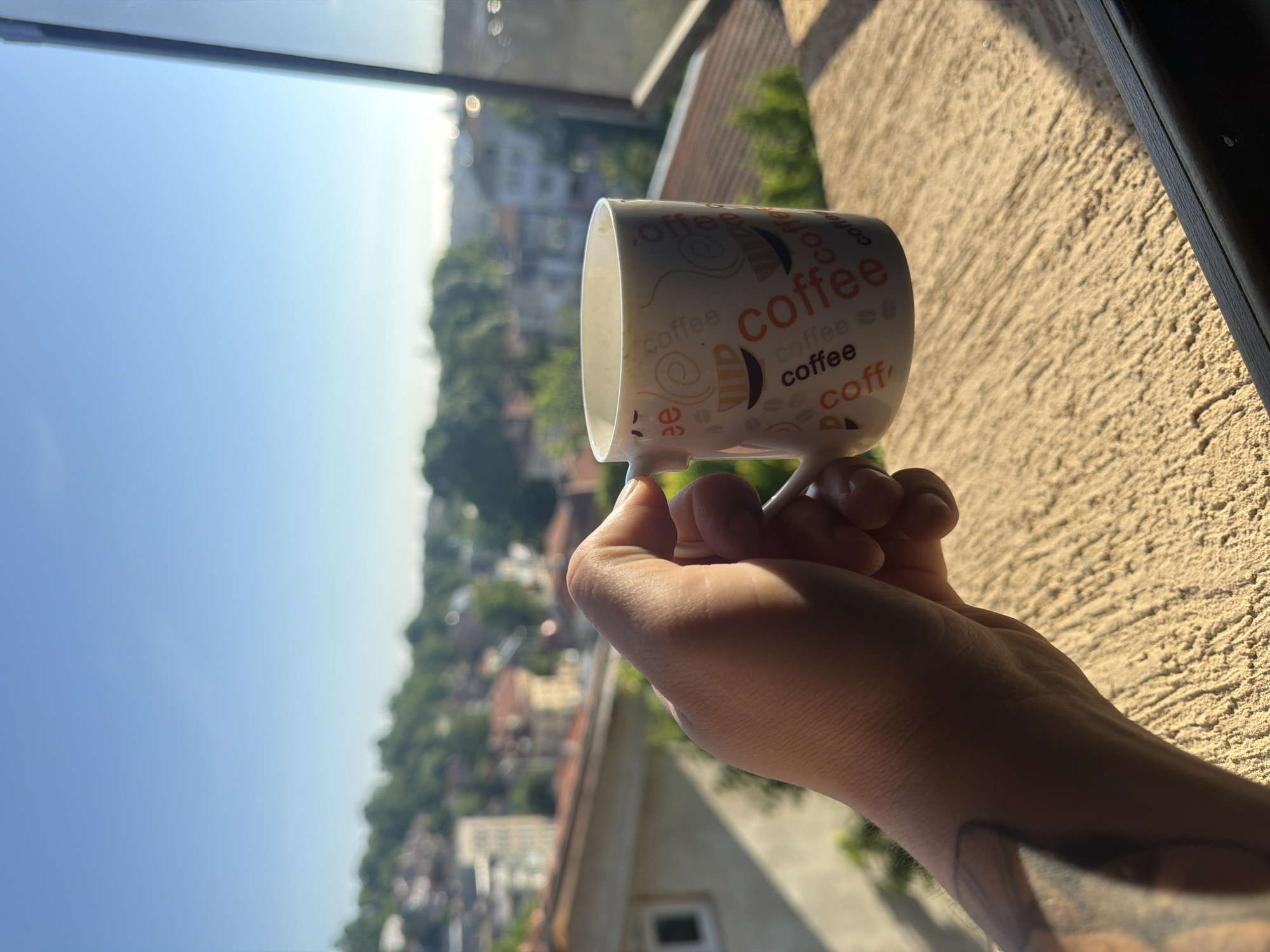 The image, presented in a vertical orientation, captures a close-up view of a person's hand holding a coffee mug against a bright, sunlit outdoor backdrop.
**Subjects:** The primary subjects are a human hand, appearing to be a right hand, and a white ceramic coffee mug. The hand has a tattoo visible on the wrist/forearm. The mug is adorned with repeated instances of the word "coffee" in different fonts and colors (notably orange and black), along with coffee bean and steam swirl motifs.
**Scene/Setting:** The background features a vibrant landscape viewed from an elevated position, likely a balcony or window. A clear, expansive blue sky occupies the upper portion of the scene. Below, a sprawling cityscape or town, consistent with Shumen, Bulgaria, is visible, characterized by numerous green trees, residential buildings with European-style architecture, and distant structures fading into a soft blur. A textured, light-colored wall with strong shadows is prominent on the right side of the frame, hinting at a building exterior, while a dark window or balcony frame is partially visible at the top and far right.
**Activity or Event:** The image suggests a peaceful moment of a person enjoying a hot beverage, likely coffee, while taking in the panoramic view of the surroundings.
**Notable Details:** The bright sunlight, casting distinct shadows on the hand and the wall, indicates a clear, sunny day, probably during the morning or early afternoon. The deep blue of the sky reinforces the notion of good weather. The focus is sharply on the hand and mug, with the background intentionally blurred to emphasize the subject.
**Visible Text:** The word "coffee" is clearly visible on the mug's surface in multiple stylized renditions.