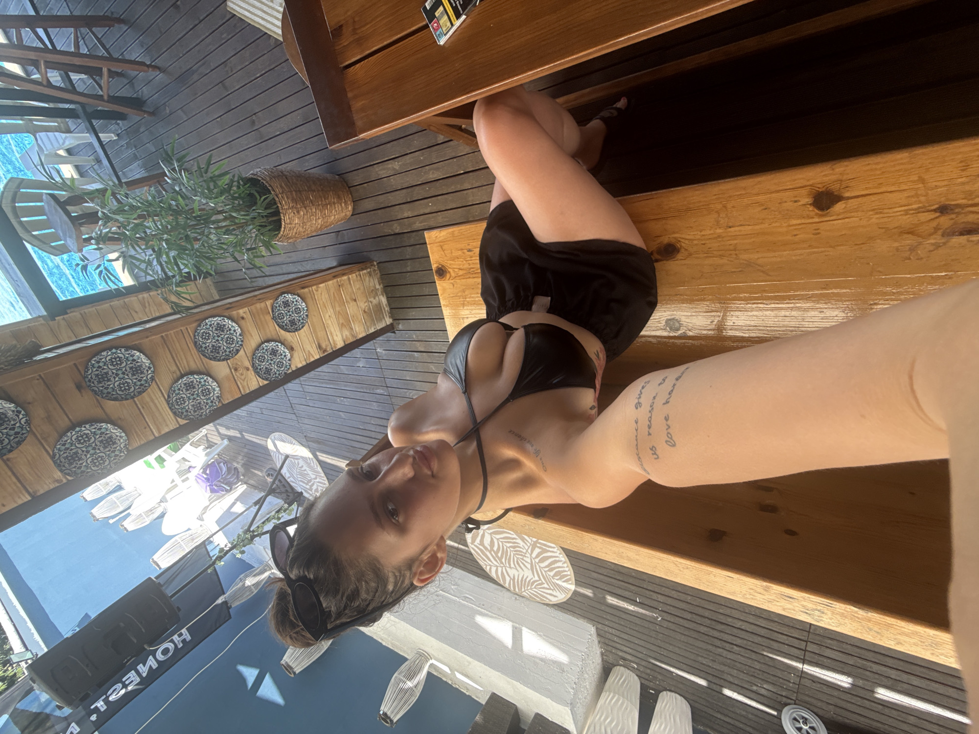 In the city of Varvara, Bulgaria, a young woman is captured in a selfie while sitting on a wooden bench. She has her hair up in a bun and is wearing a black bikini top and a black skirt. Her arm, which is holding the phone, has a tattoo that reads, "distance gives us reason to love harder." The background shows a blue pool and wooden decking, suggesting an outdoor setting perhaps a beach club or resort. The woman appears relaxed and is looking towards the camera.