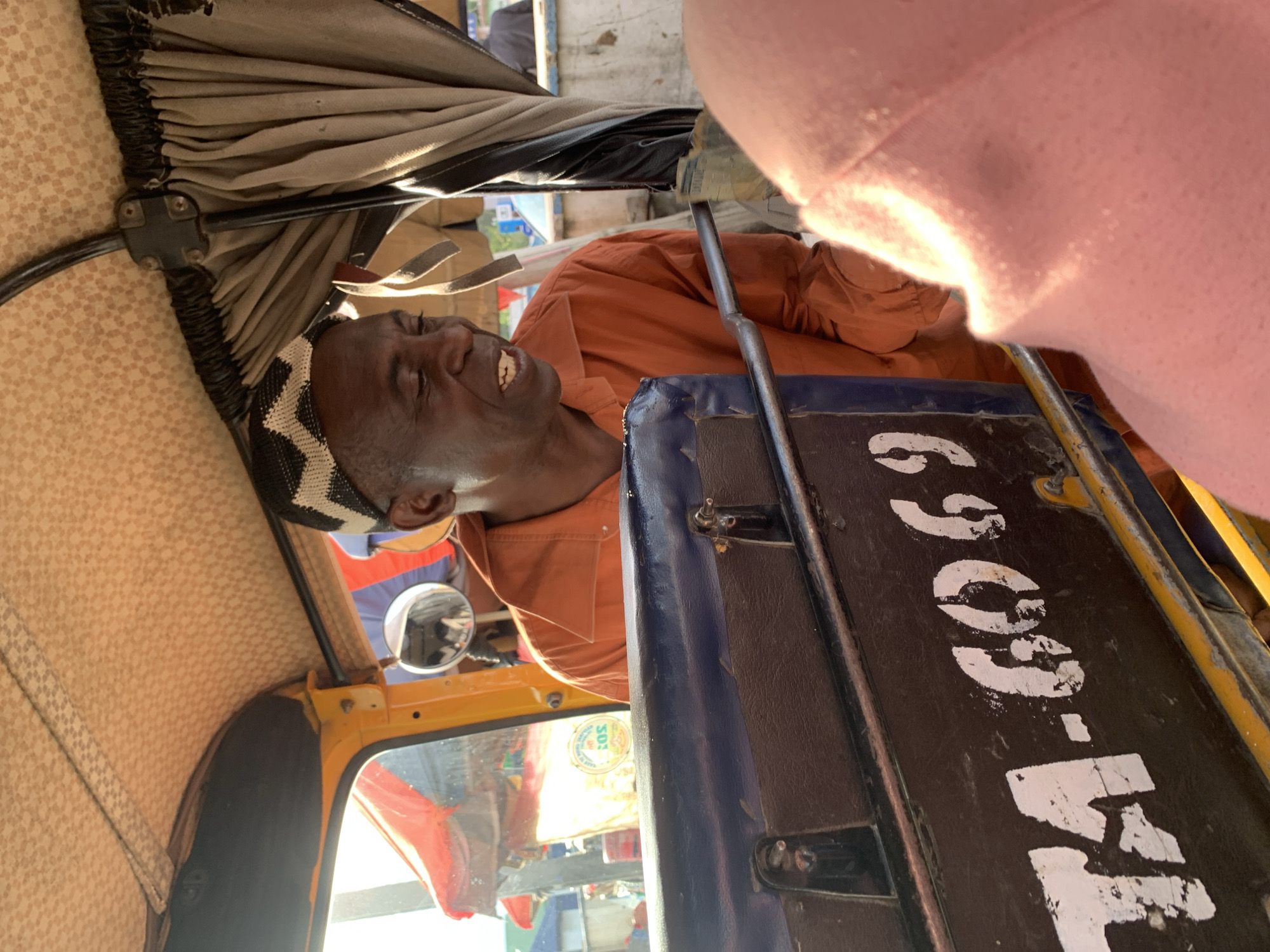 In a brightly lit outdoor environment, an elderly African man, wearing an orange collared shirt and a black and white zig-zag patterned cap, is seated inside a yellow tuk-tuk. He is looking to his right, with his mouth slightly open as if speaking. The tuk-tuk's interior features a patterned fabric lining on the ceiling and tan curtains on the right side. A blue and black dashboard with white lettering, including "TA-6069", is visible in the foreground, partially obscuring the man. The backdrop shows glimpses of the bustling city of Jos, Nigeria, with hints of colorful awnings and other vehicles. The overall scene captures a moment of everyday life within the city.