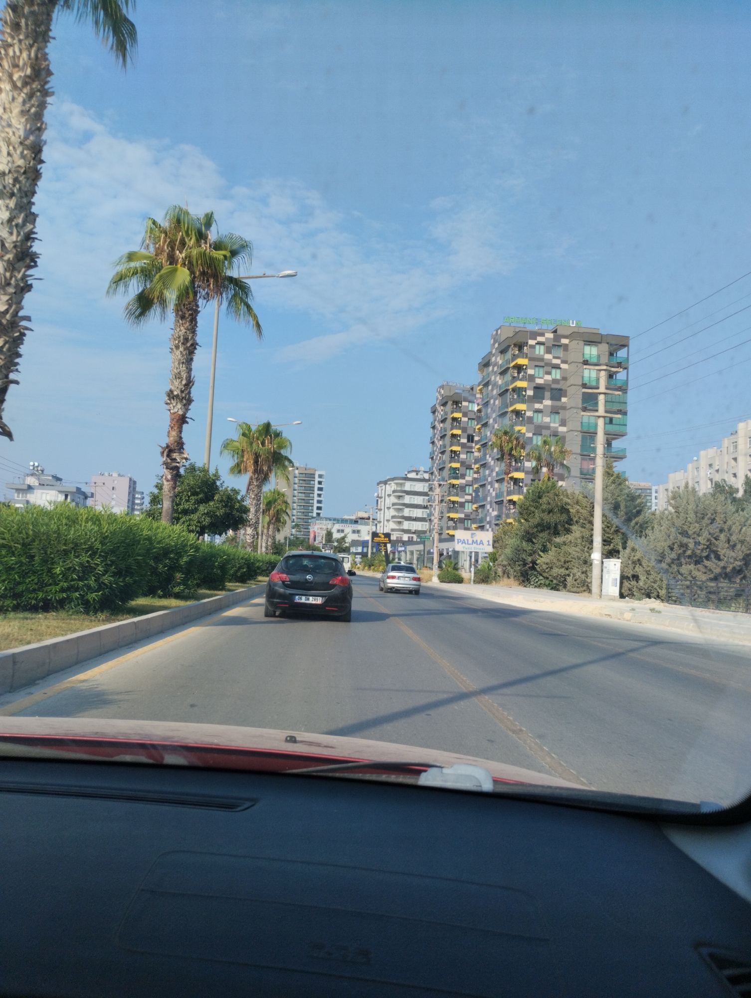 The image captures a street scene in Mezitli, Turkey, on a clear, sunny day. A vehicle, likely a hatchback, is seen driving down a multi-lane road. Palm trees line the roadside, with dense green bushes and some trees also present. In the background, modern apartment buildings, some with balconies and a distinct grey and yellow facade, dominate the skyline. Power lines are visible overhead. The perspective is from inside another vehicle, looking forward through the windshield. The dashboard of the vehicle is partially visible in the foreground. The road surface is asphalt with visible lane markings.