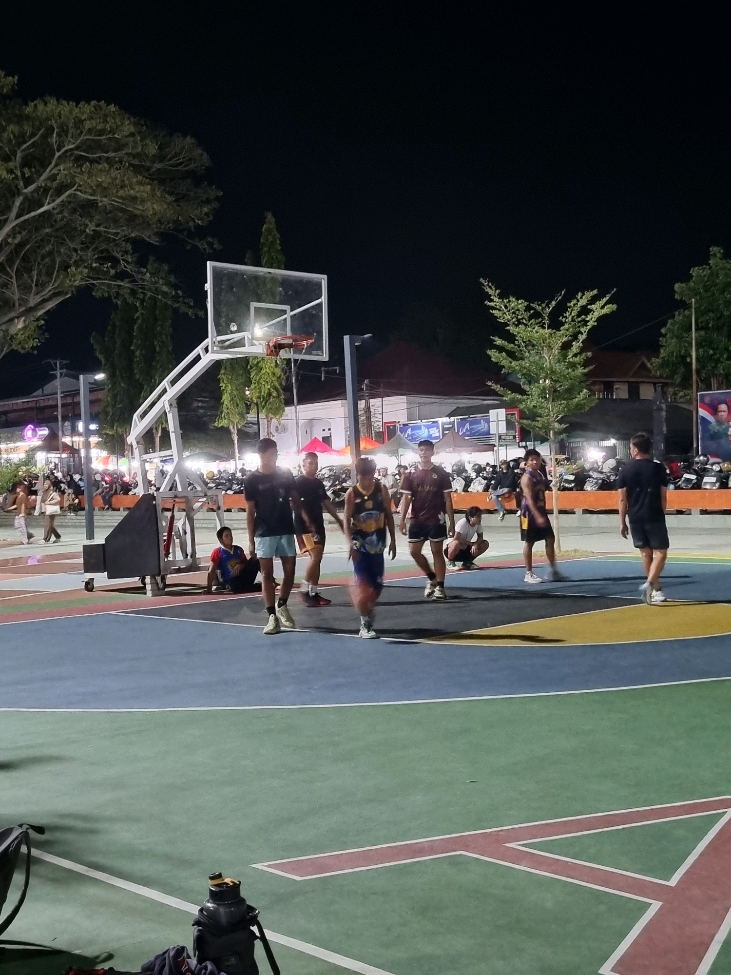 A group of young men are playing basketball on an outdoor court at night in Tanamodindi, Indonesia. The court is illuminated by artificial lighting. Some players are in motion, while others are standing or observing. The court has a multi-colored surface with blue, green, and yellow sections. In the background, there are trees, buildings, parked motorcycles, and a crowd of spectators. A basketball hoop with a glass backboard is visible, along with a portable base. One person is sitting on the ground near the hoop. A bag and a water bottle are on the green portion of the court in the foreground. The scene appears to be lively and recreational.