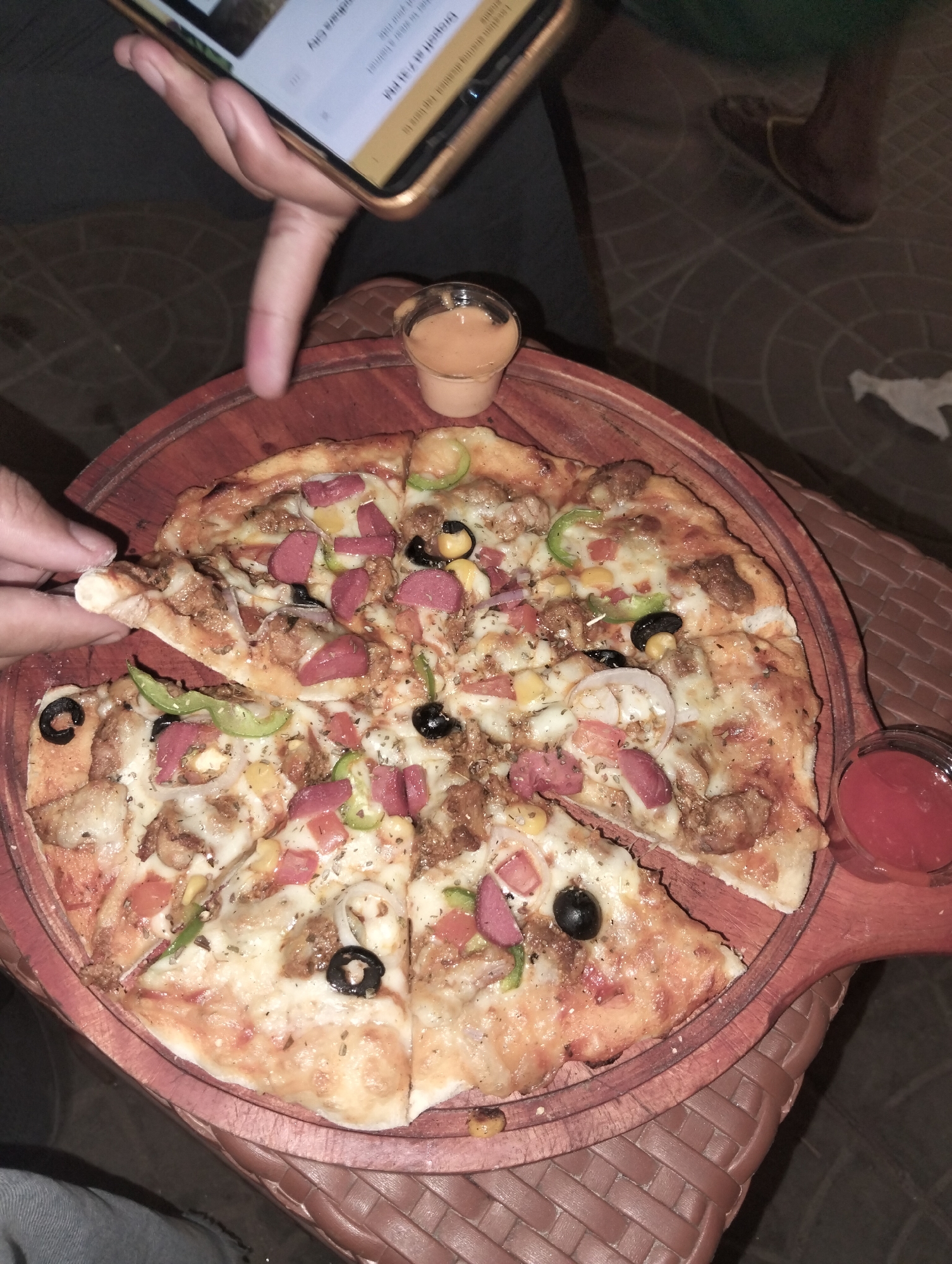 Here is a description of the image:
A person is taking a slice of pizza from a wooden platter. The pizza is topped with cheese, meat, olives, corn, tomatoes, onions, and green peppers. Two small cups of sauce are placed on the edges of the platter. The pizza and platter are on top of a brown woven basket. In the background, a person is holding a phone, and shoes can be seen on the floor. Based on the OCR text detected in the image, this image was taken in Dhaka city in Bangladesh.