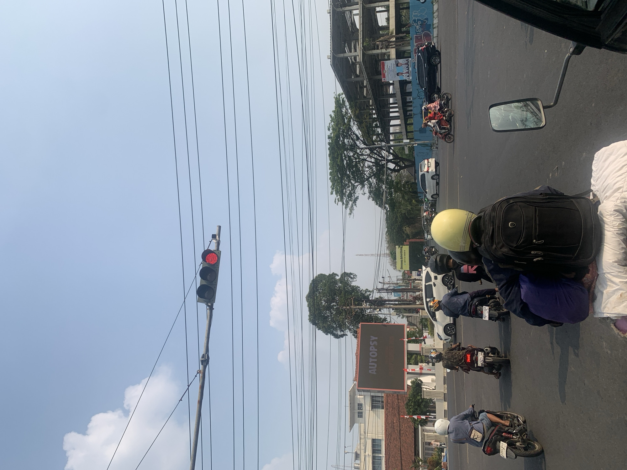 In a city intersection in Palembang, Indonesia, a traffic light displays a red signal, causing a queue of motorcycles and cars to halt. The foreground is dominated by the back of a motorcycle and its rider, distinguished by a yellow helmet and a black backpack. A street sign that reads "Demang Advertising" is visible in the distance, accompanied by a large digital billboard displaying the word "AUTOPSY". The intersection is bordered by buildings, including a white structure with red-tiled roofs and a partially constructed building. Several trees punctuate the urban landscape, and a network of power lines crisscrosses the sky. The overall atmosphere suggests a typical daytime scene in an urban environment.