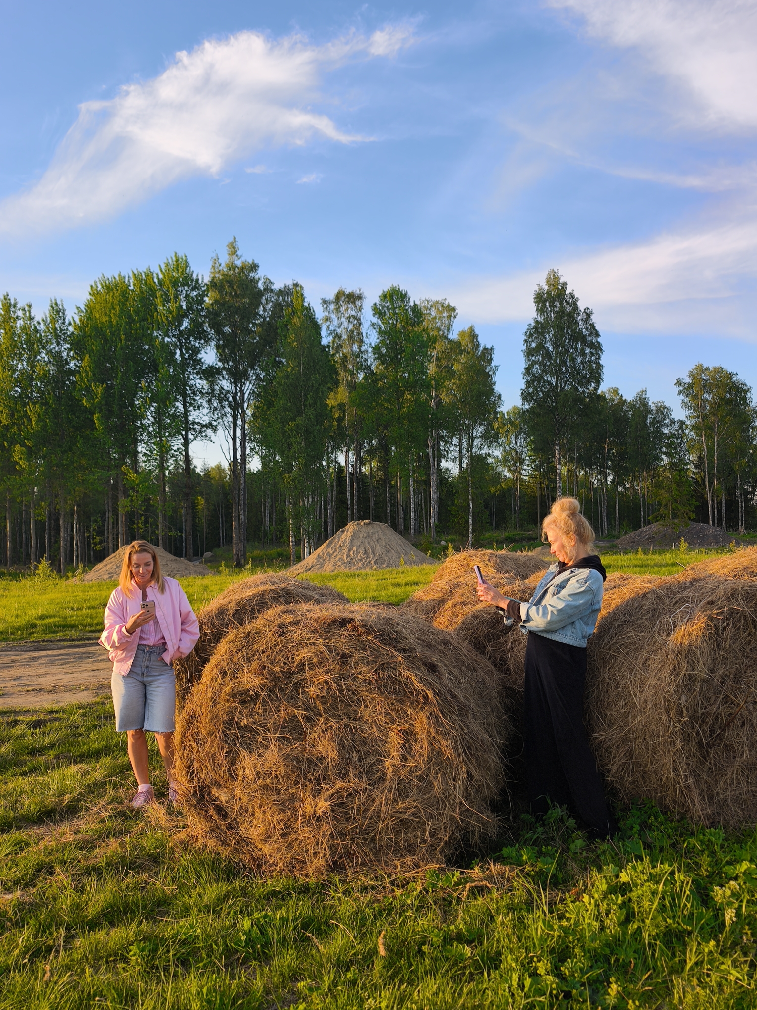 This image captures an outdoor scene in Стеклянный, Russia, during the warm glow of late afternoon or early evening, indicated by the golden light illuminating the landscape. Two women are present in a grassy field, surrounded by several large, round hay bales.
On the left, a woman with blond hair, wearing a light pink jacket over a striped shirt, light denim shorts, and pink sneakers, stands in a relaxed posture. She is holding a smartphone in her hands, looking down at its screen. On the right, another woman, also with blond hair styled in a bun, is dressed in a light blue denim jacket over a dark top and a long black skirt or dress. She stands beside a hay bale, holding her smartphone up as if taking a photograph or video.
The background features a dense line of tall, slender deciduous trees, forming a natural forest edge. Further back, several large piles of sand or earth are visible. The ground is a mix of green grass and a lighter dirt path. The sky is bright blue with scattered, wispy white clouds. The overall impression is one of casual leisure in a rural, natural setting, with the subjects engaged with their digital devices. No visible text is present in the image.