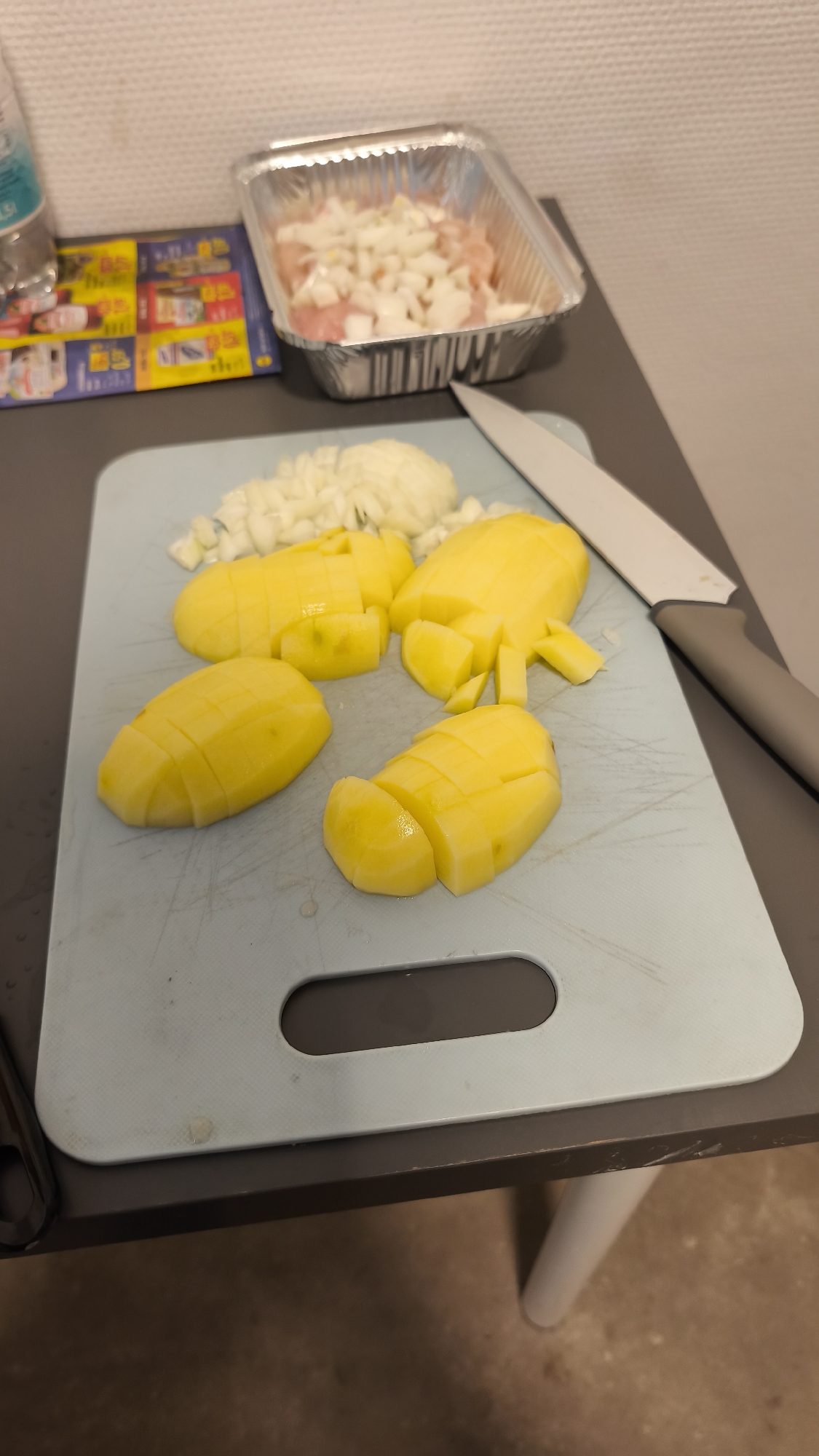 The image displays a close-up scene of food preparation taking place on a dark grey table surface, likely in a kitchen environment. The central subject is a light blue cutting board, which holds several peeled yellow potatoes that have been roughly chopped into large, irregular pieces. Next to the potatoes, a mound of finely diced white onion is visible. A kitchen knife with a grey handle and a white blade rests on the right edge of the cutting board.
In the background, positioned on the tabletop, an aluminum foil container holds raw, diced meat, which appears to be chicken, generously covered with chopped white onion. To the left of the foil container, a colorful, folded brochure or flyer featuring small images of various food products and prices is laid out. Behind the flyer, a clear plastic bottle is partially visible, with the letters "LSI" discernable on its label. The background wall is light-colored and exhibits a subtle, textured pattern. The scene depicts an active stage of preparing ingredients for a meal. No people are visible, and no specific time of day or emotions can be inferred.
