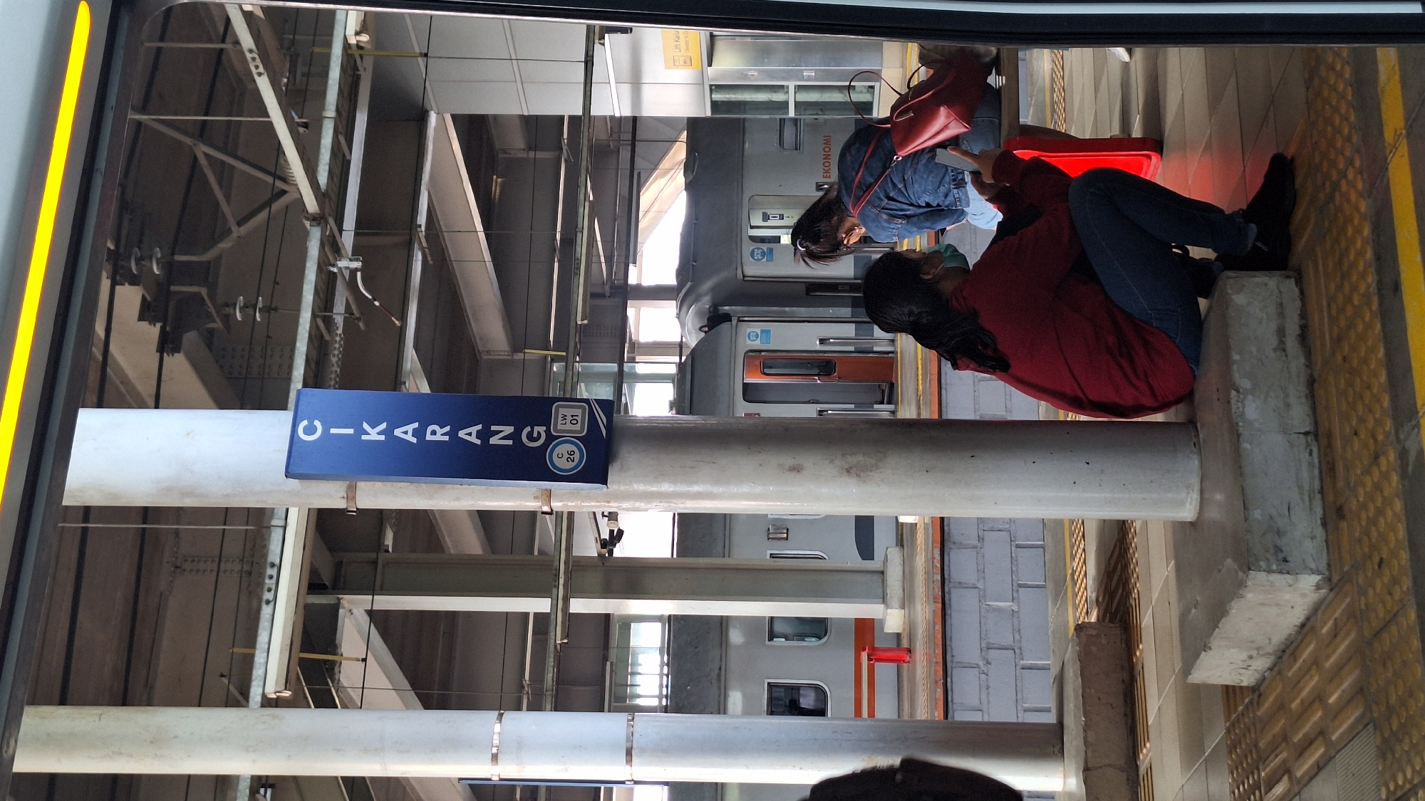 The image depicts a train station platform in Cikarang, Indonesia. A tall, blue sign with white lettering reads "CIKARANG" vertically. Below the name, there are two circular emblems, one with "C 26" and the other with "LW 01". Several thick, cylindrical pillars support the station's roof structure. In the foreground, a person wearing a red hoodie and jeans squats down, looking at their phone. They are wearing a light blue face mask. To their right, another person in a denim jacket and jeans, also wearing a face mask, is partially visible. They are holding a smartphone. A red backpack rests on a bench behind them. A train with gray carriages and orange doors is stopped at the platform, with the word "EKONOMI" visible on one of the carriages. The platform has tactile paving and yellow lines marking the edge. The environment suggests an active transit hub.