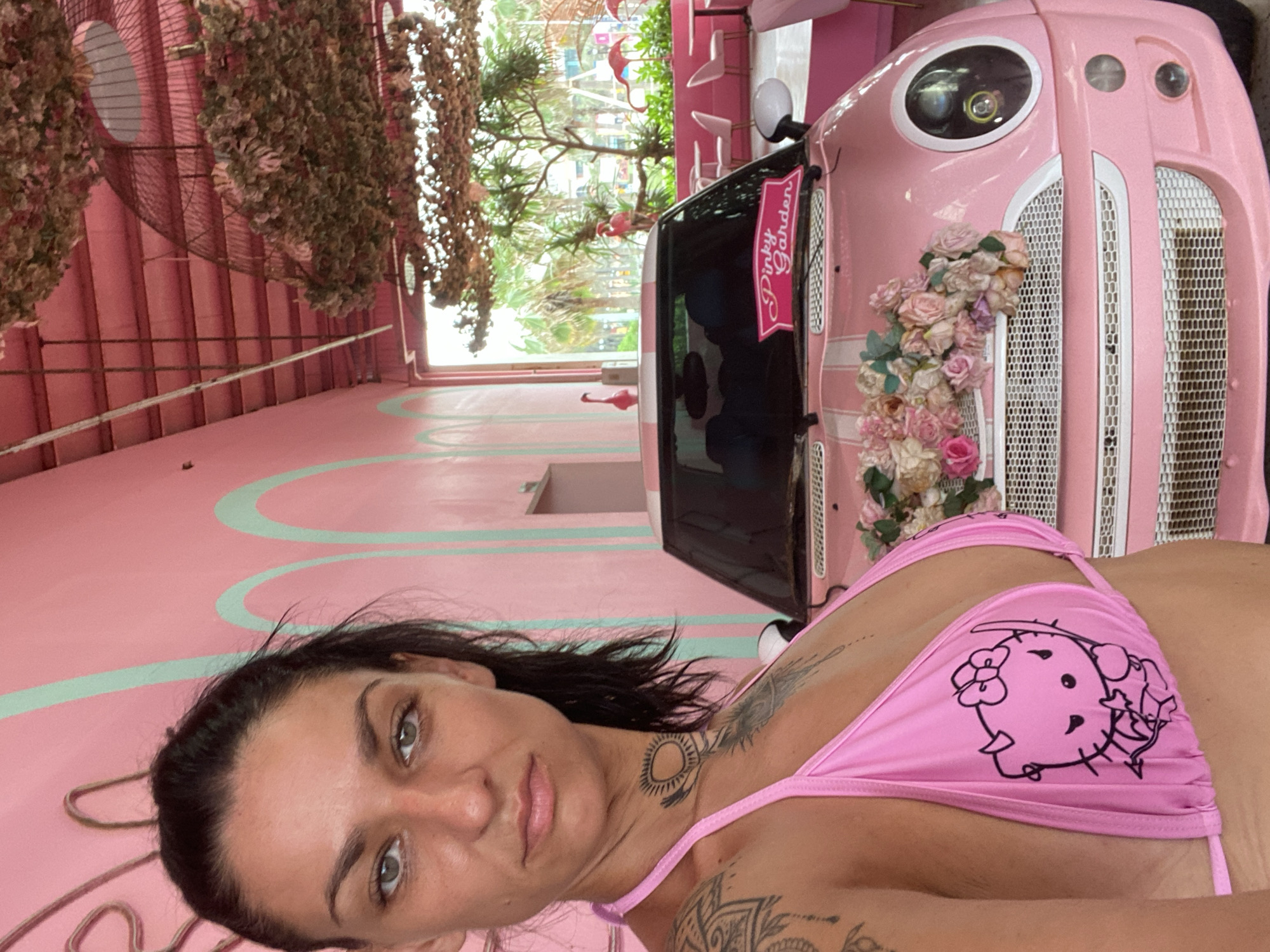 The image presents a selfie-style portrait of a woman in a vibrantly pink-themed environment, likely a cafe or attraction, set in a daylight scene. The woman, positioned in the foreground and looking directly at the viewer, has dark hair, light eyes, and a neutral expression. She wears a bright pink bikini top featuring a black outline drawing of the Hello Kitty character. Visible tattoos include a circular sun-like design on her neck and a mandala-like pattern on her shoulder.
Behind the woman, to her right, is a custom pink vehicle resembling a small retro car or golf cart. It has a prominent round headlight, a white-trimmed grille, and is adorned with clusters of artificial roses in shades of pink, white, and cream. A banner on the side of the vehicle clearly displays the text "Pinky Garden." The surrounding setting is characterized by entirely pink walls, and the floor features decorative light blue wavy lines. Overhead, strands of dried or artificial foliage hang down as part of the decor. In the background, through an open space, green trees and several pink flamingo figurines are visible, reinforcing the whimsical, garden-like theme suggested by the "Pinky Garden" name. The overall atmosphere is playful and decorative, consistent with themed establishments found in locations like Tien Thanh Ward, Vietnam.