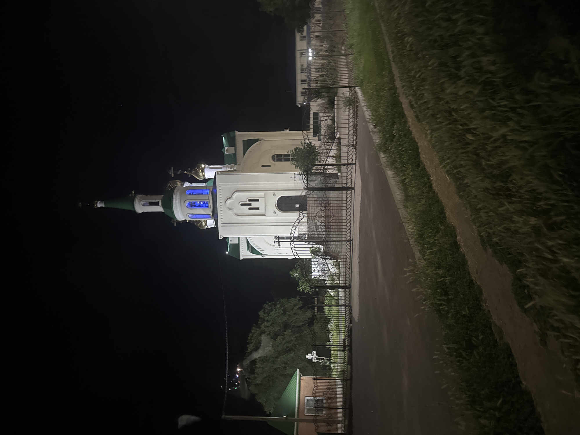 The image displays an Eastern Orthodox church at night, illuminated by artificial lights. The church is white with green and gold domes. Blue lights shine from some of the windows, creating a striking contrast with the dark sky. A black metal fence encloses part of the church grounds. To the right of the church, there is a grassy area and a dirt path, suggesting a rural or semi-rural setting. In the background, other buildings are visible, also illuminated.
The scene is set at night, as indicated by the dark sky and the artificial lighting on the structures. The presence of grass and a path points to an outdoor location. The overall mood is serene and perhaps a bit majestic due to the illuminated architecture against the night sky. There are no people visible in the image. The specific location is Yegorlykskaya, Russia, though this context is not visually evident within the photograph itself. No visible text is present.