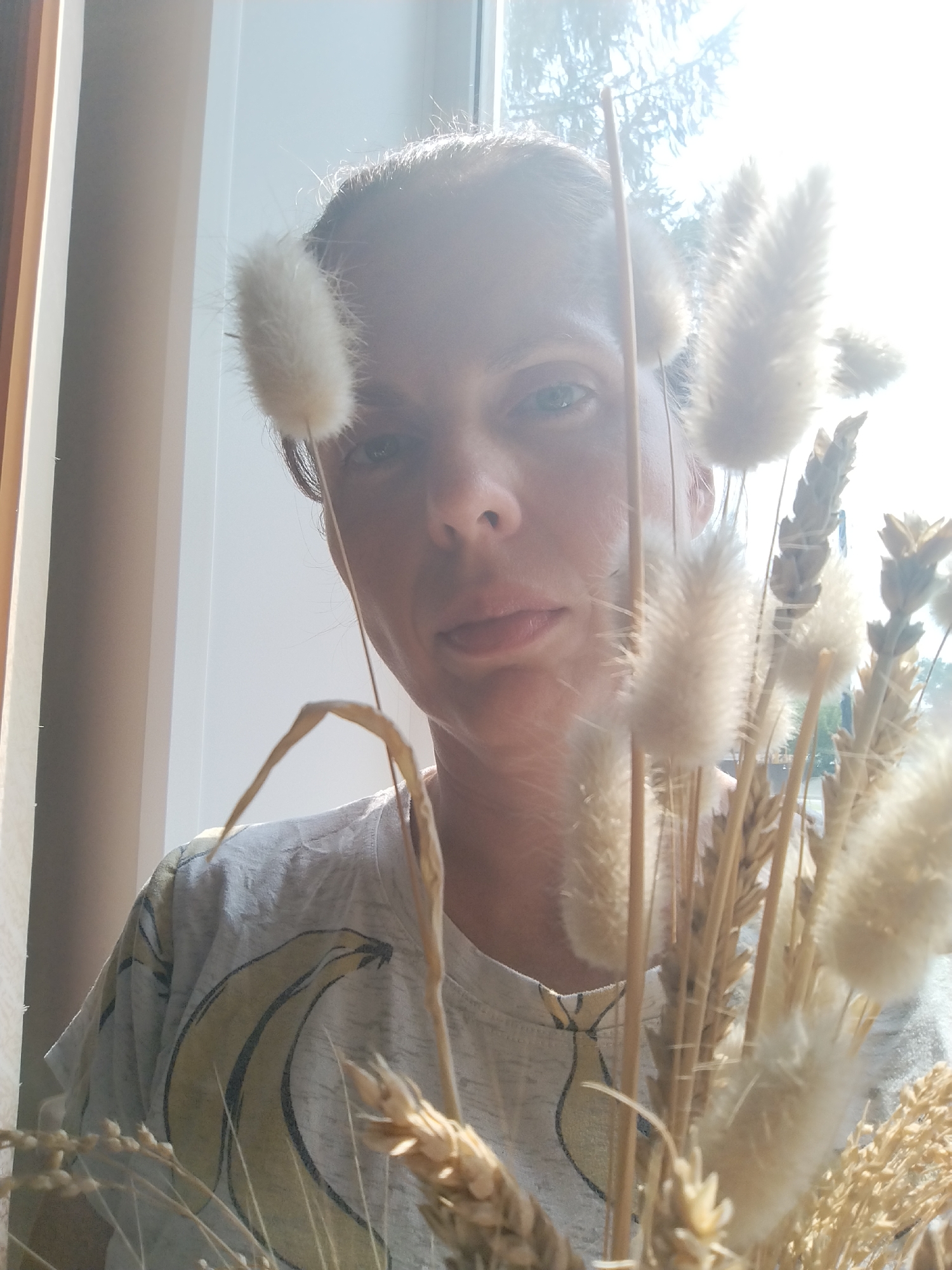 A selfie, taken from a close-up perspective, shows a person holding a bouquet of dried flowers and wheat stalks in front of their face. The person is wearing a light-colored t-shirt with a graphic of two bananas. The background is out of focus, with a window providing bright natural light, suggesting an indoor setting near a window. The scene is filmed in Zavodoukovsk, Russia.