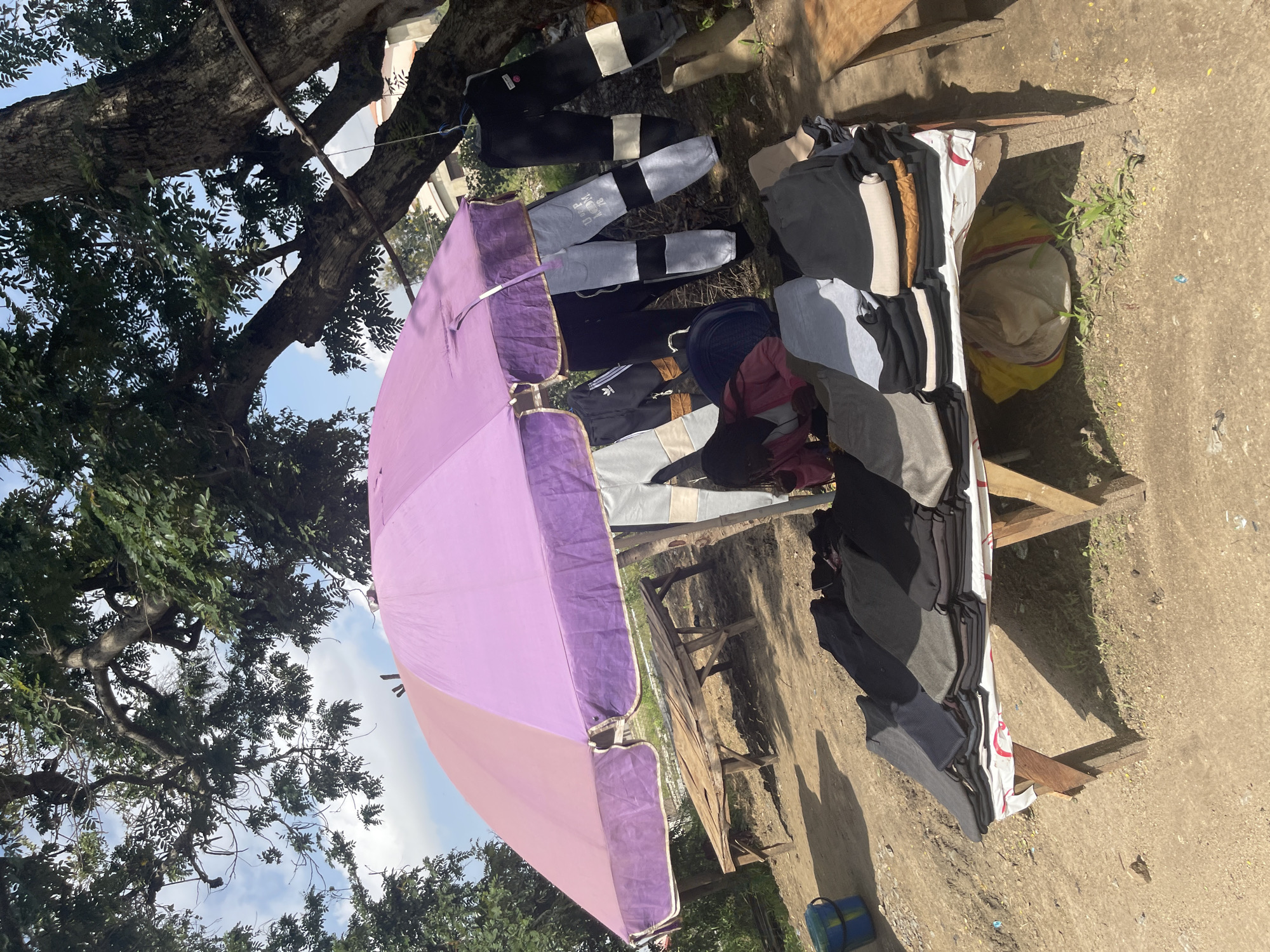 A vendor displays clothing on a table under a large, purple umbrella outdoors in Rafin Par, Nigeria. The table is laden with folded garments, predominantly black and grey trousers, shirts, and some other items of clothing. To the left of the table, a large tree with lush green foliage shades the scene. The ground is a dusty, earthy texture with some patches of dry grass. A wooden bench is visible behind the table, and further back, there's a hint of a building. Sunlight casts shadows from the table and the tree, suggesting it is daytime, possibly mid-morning or afternoon. The overall atmosphere appears to be that of a typical street market in Nigeria, with the vendor setting up for business. The crops highlight the variety of clothing, the textures of the tree bark and leaves, and the ground underfoot. The image is oriented vertically, with the sky visible at the top left and the ground at the bottom right.
