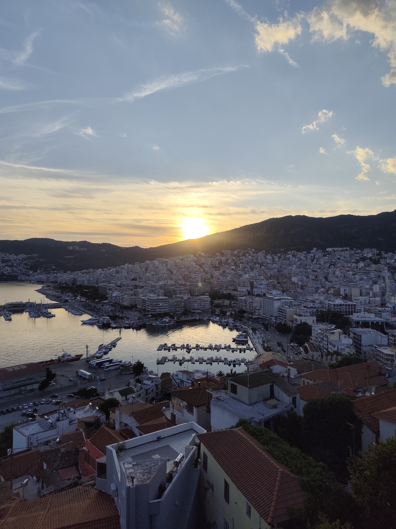 This image captures a scenic sunset over the coastal city of Kavala, Greece. The sun is low on the horizon, casting a warm, golden glow across the sky and reflecting on the calm waters of the harbor. Wispy clouds, tinged with orange and yellow, are scattered across a pale blue sky, indicating fair weather.
The foreground shows a densely packed urban landscape with buildings featuring terracotta-tiled roofs, characteristic of Mediterranean architecture. The cityscape extends towards the harbor, where numerous boats and yachts are docked, their masts creating a delicate silhouette against the water. A large red vessel is prominently visible in the lower left of the frame. Beyond the city, a dark, tree-covered hill forms the backdrop, its slopes dotted with more buildings, suggesting the city's expansion upwards.
The overall scene evokes a sense of tranquility and beauty, typical of a peaceful evening in a coastal town. The light suggests it is late afternoon or early evening. No people are clearly discernible in the image, and there is no visible text. The view is from an elevated position, offering a panoramic perspective of the harbor, the city, and the surrounding natural landscape.