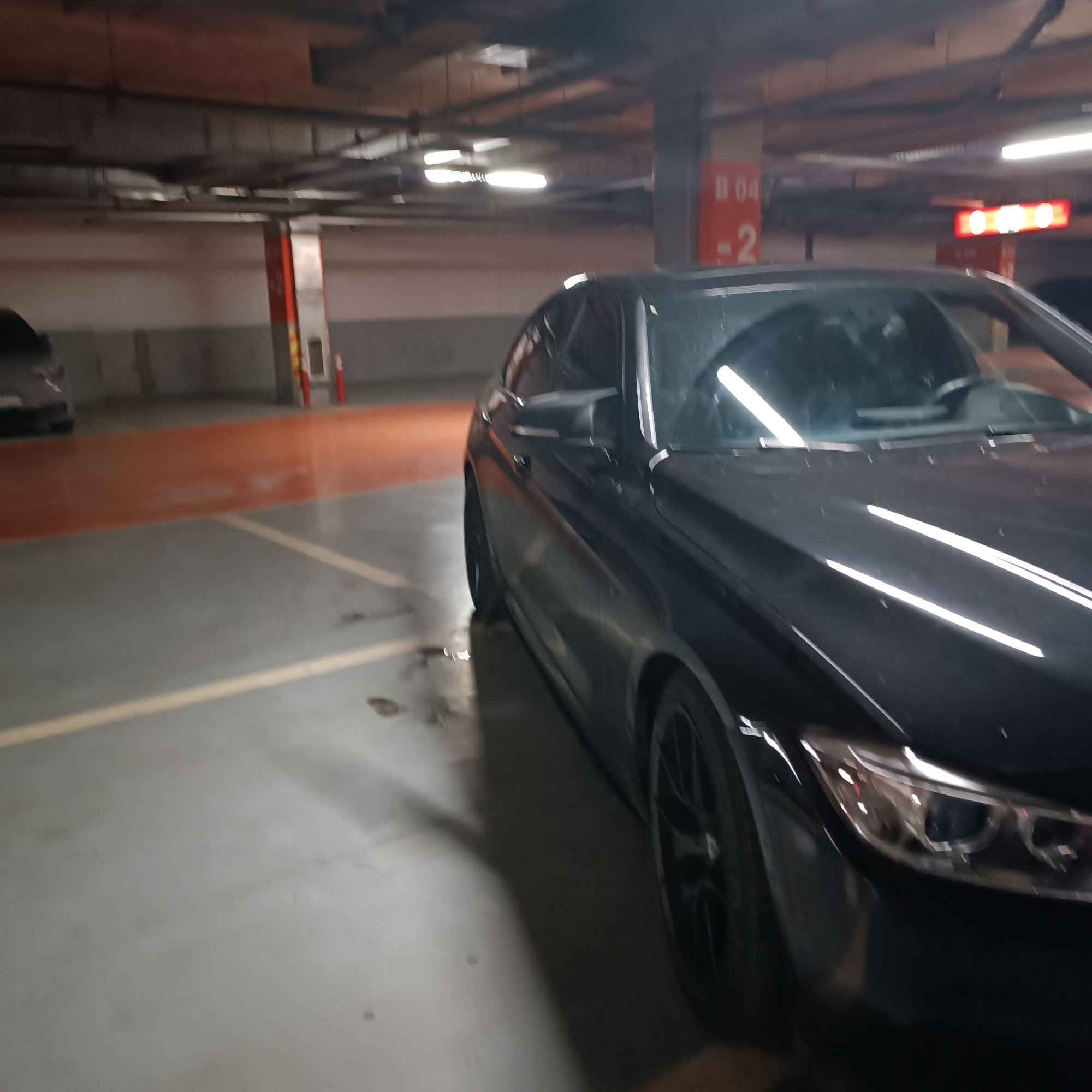 The image captures a black car parked in an indoor parking garage in Sofia, Bulgaria. The car, appearing to be a sedan or coupe, occupies the right side of the frame, with its front right headlight, wheel, and passenger side visible. The car's glossy black finish reflects the bright, artificial overhead lighting.
In the background, a lighter-colored car is partially visible on the far left. The parking environment features concrete pillars, exposed pipes, and ventilation ducts across the ceiling. The grey concrete floor is marked with painted white or yellow parking lines, one of which runs prominently in the foreground. Noticeable wet patches or water stains are present on the floor near the front right tire of the black car.
A red sign is attached to a pillar in the mid-ground, clearly displaying the text "B 04" at the top and "-2" beneath it, likely indicating a section and level within the garage. Further in the distance, another red, illuminated sign is visible, but its text is indistinguishable. Red and white safety bollards or cones are also discernible next to a pillar in the background. The scene is well-lit by fluorescent lamps, typical of an underground or multi-story parking facility. No people are visible, and no specific activity is taking place other than the cars being parked.