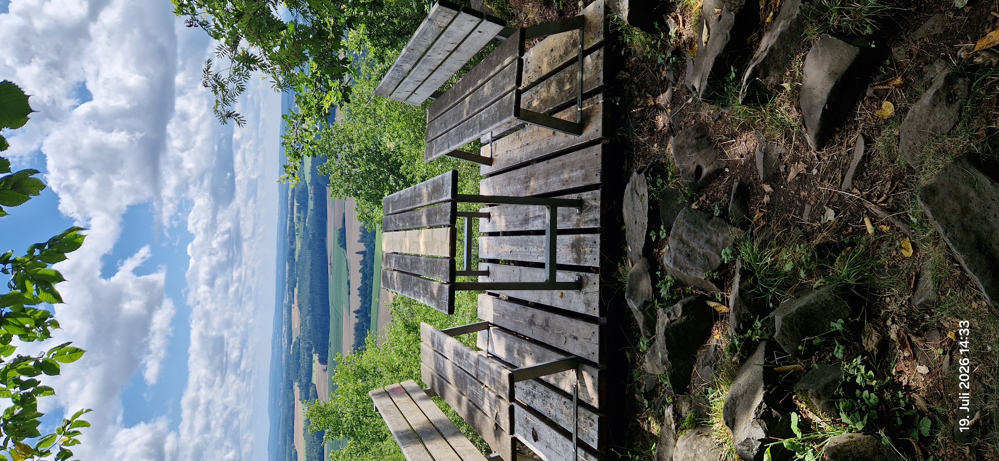 This image shows a wooden picnic table and benches with a scenic view of rolling hills and fields under a partly cloudy sky. The foreground is a rocky, wooded area. The date and time stamp "19. Juli 2026 14:33" is visible in the bottom left corner. The location is identified as Neustadt am Kulm, Germany, suggesting this is a viewpoint or rest area in a natural setting. There are no people present in the image.