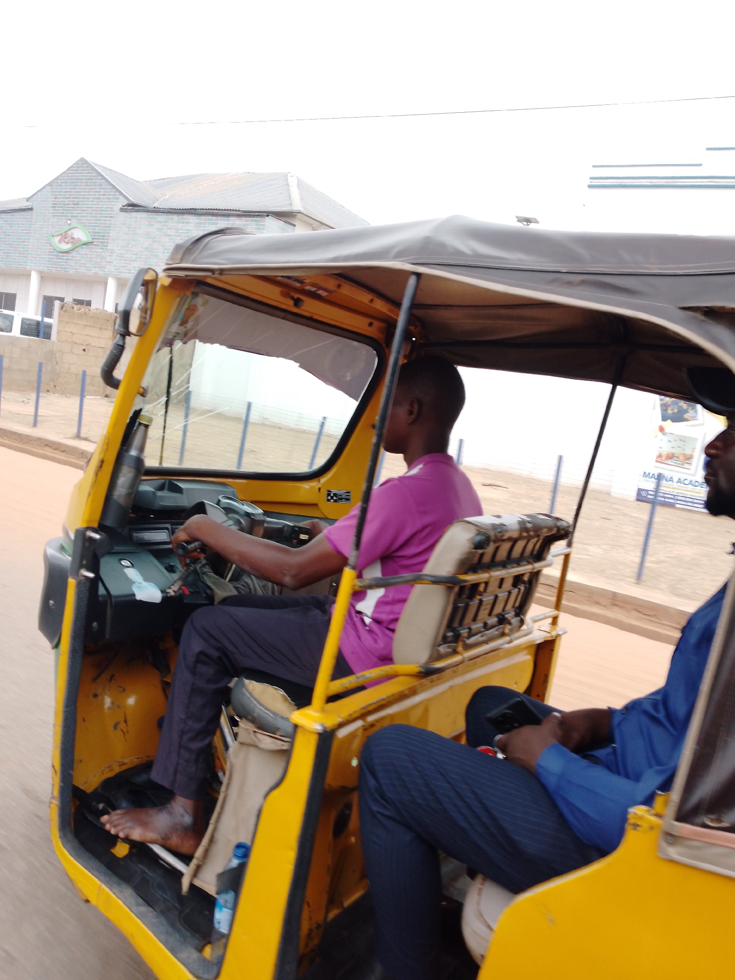 A yellow "keke" (auto rickshaw) is being driven along a road in Sabon Tasha, Nigeria. The driver, wearing a pink shirt and dark pants, is barefoot. A passenger, wearing a blue shirt and pinstripe pants, is seated in the back and holding a dark-colored smartphone. The background shows a building and a sign that reads "MAINA ACADEMY." The road appears to be dusty.