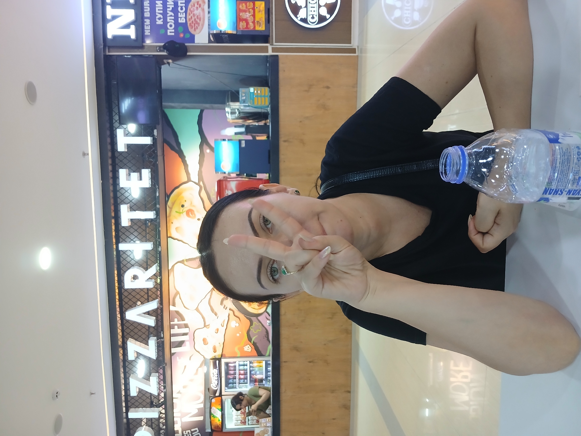 This media file shows a portrait of a woman in a brightly lit indoor setting, likely a mall food court or similar commercial establishment. The woman is positioned in the foreground, her body angled towards the camera. She is wearing a black t-shirt and has long, dark hair. Her left hand is raised, forming a peace sign with her index and middle fingers, and she is looking directly at the camera with a relaxed expression. A bottle of water rests on the table beside her.
Behind the woman, a food establishment with prominent signage is visible. The signage includes the words "PIZZARTE" and "CHICKEN" in large letters. The establishment appears to have colorful displays and what looks like refrigeration units, possibly for drinks and food items. Reflective surfaces and overhead lighting suggest an indoor environment. Text in Cyrillic script is also visible on some of the signage, including "НОВЫЙ БУРГЕР", "КУПИ", "ПОЛУЧИ", and "БЕСПЛАТНО", indicating a Russian-speaking region. The overall atmosphere appears casual and bustling. The image is oriented vertically.