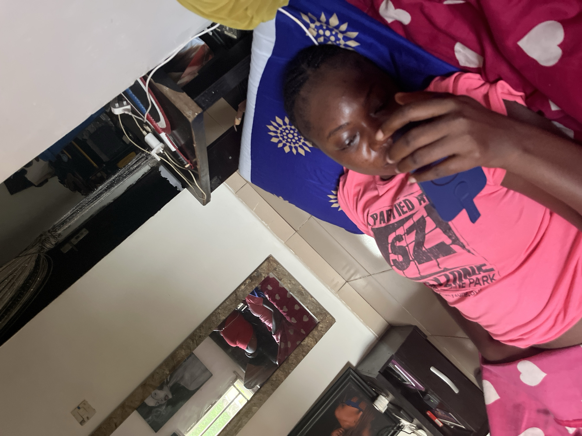 The image depicts an indoor scene, likely a bedroom in Abuja, Nigeria, with a young black individual lying on a bed. The person, who appears to be female, is wearing a bright pink t-shirt and pink shorts or is covered by a pink blanket adorned with white heart patterns. They are resting their head on a blue pillow featuring a sunburst design and are engaged with a mobile phone in a blue case, holding it with their left hand while their right hand is near their face. Their expression is neutral, suggesting relaxation.
The pink t-shirt has visible text, partially decipherable as "PARTIED A" above a large "SZ" or "5Z" and then "ZONE PARK" with smaller, unclear text below.
To the left of the bed, a collection of white power adapters and tangled wires is visible, connected to a power strip or outlet, suggesting various devices are being charged or powered. These plugs appear to be of the Type G (UK/Nigerian) standard. Adjacent to the wiring is a dark wooden structure, possibly a bedside table or shelf. The floor is covered with light-colored square tiles.
In the background, a rectangular mirror with a textured frame is mounted on the wall, reflecting an outdoor view through a window, showing green foliage and daylight. Below the mirror, a framed picture featuring a black and white portrait or figure is displayed. To the right of the person's lower body, a dark brown cabinet or chest of drawers with a silver handle is partially visible. The overall setting suggests a private living space during the daytime.