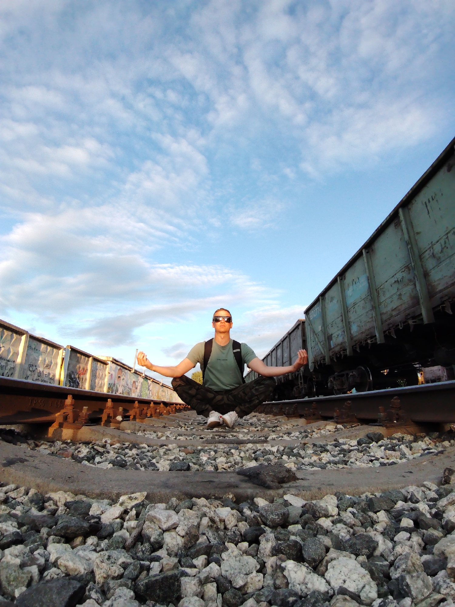 An adult male sits cross-legged in a meditative posture on the gravel ballast between two sets of parallel railroad tracks in Chaikovsky, Russia. He wears dark sunglasses, an olive-green t-shirt with a small graphic, camouflage pants, and white sneakers, with a dark backpack on his shoulders. His hands are open with palms facing upward, and his thumbs and index fingers touch in a mudra gesture. To the left, a line of stationary freight cars with colorful graffiti on their sides runs parallel to the tracks. To the right, another line of weathered, dull blue-green freight cars with visible rust and faded stenciling also runs parallel. The foreground consists of densely packed gray and white gravel. The scene is illuminated by natural light under a partly cloudy, light blue sky with white, streaky clouds, indicating either early morning or late afternoon.