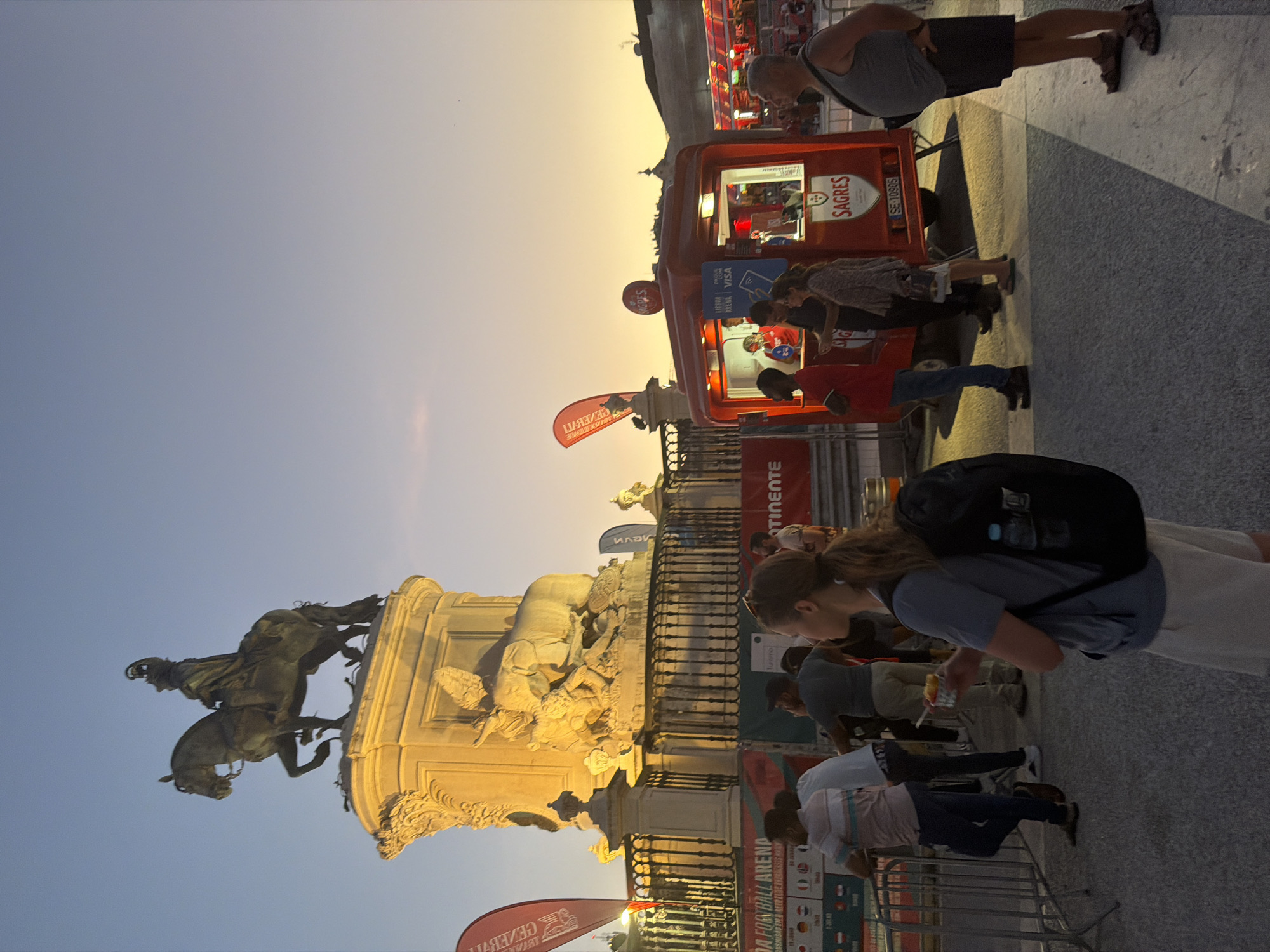 The image depicts an outdoor scene in Lisbon, Portugal, at dusk, likely near a public square or monument. In the background, a large equestrian statue stands atop a monumental pedestal adorned with sculptures of elephants and other figures. The statue and pedestal are illuminated by artificial lights, casting a warm glow against the darkening sky.
In the middle ground, a red food truck with "SAGRES" branding is parked, serving customers. Several people are gathered around the truck, some waiting in line and others receiving food or drinks. The truck has a service window where individuals are visible preparing orders. Banners and flags are present, including a red flag with a silhouette of a person on a horse, and another red flag with text.
In the foreground, a woman wearing a grey t-shirt, light-colored shorts, and a black backpack is holding a small cup of food. She is looking to her right. To her left, a metal barrier separates the public from an area behind it, where other individuals are standing. The ground is paved with cobblestones or similar material. The overall atmosphere suggests a lively event or gathering. The presence of the food truck and the crowd indicates a public space where people are congregating. The background suggests a historically significant location, likely a plaza given the prominent monument.