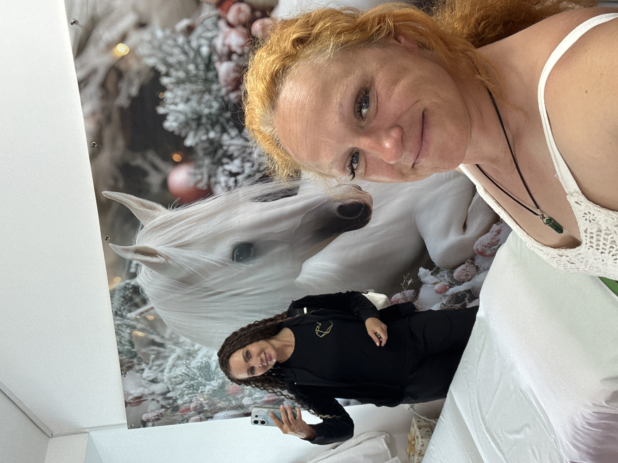 In a brightly lit room, likely a spa or treatment facility, two women are captured in a selfie. A large, vibrant mural of a white horse adorned with festive, snowy foliage and soft bokeh lighting serves as a backdrop. The woman on the left, with long, dark curly hair and wearing a black outfit, holds a smartphone to take the picture. The woman on the right, with vibrant red hair and wearing a white crocheted top, is positioned closer to the camera, smiling at the lens. In the foreground, a white massage table is visible, neatly covered with linens. The overall impression is one of a professional yet welcoming atmosphere. There is no specific indication of the location within Chaikovsky, Russia.