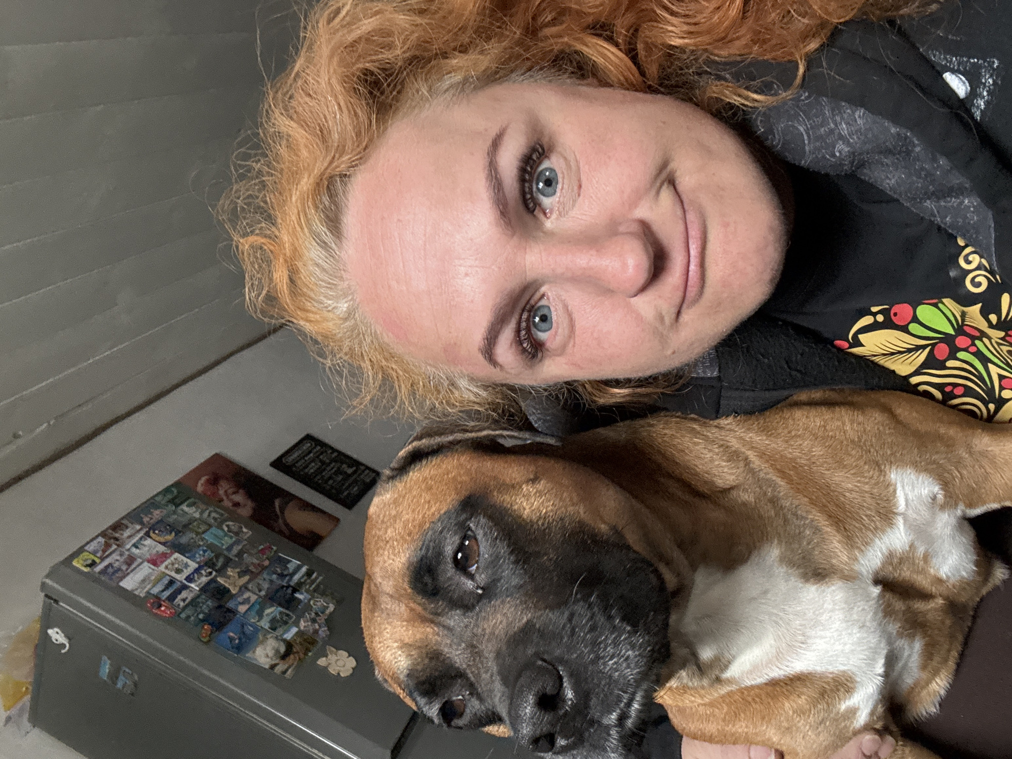 A selfie-style, vertically oriented close-up shows a woman with red, curly hair and blue eyes looking into the camera. She has fair skin and a slight smile. The woman wears a dark, patterned shirt.
Partially visible beneath her is a brown and black dog. The dog's head is tilted slightly, with one eye visible and appearing to look off to the side. The dog's fur is short and smooth.
Behind the woman and the dog, a gray refrigerator is visible. The refrigerator door is covered with small magnets. Above the refrigerator, a dark wall with a small framed object is seen.
The image appears to have been taken indoors. The location in Chaikovsky, Russia, is not possible to determine from the image.