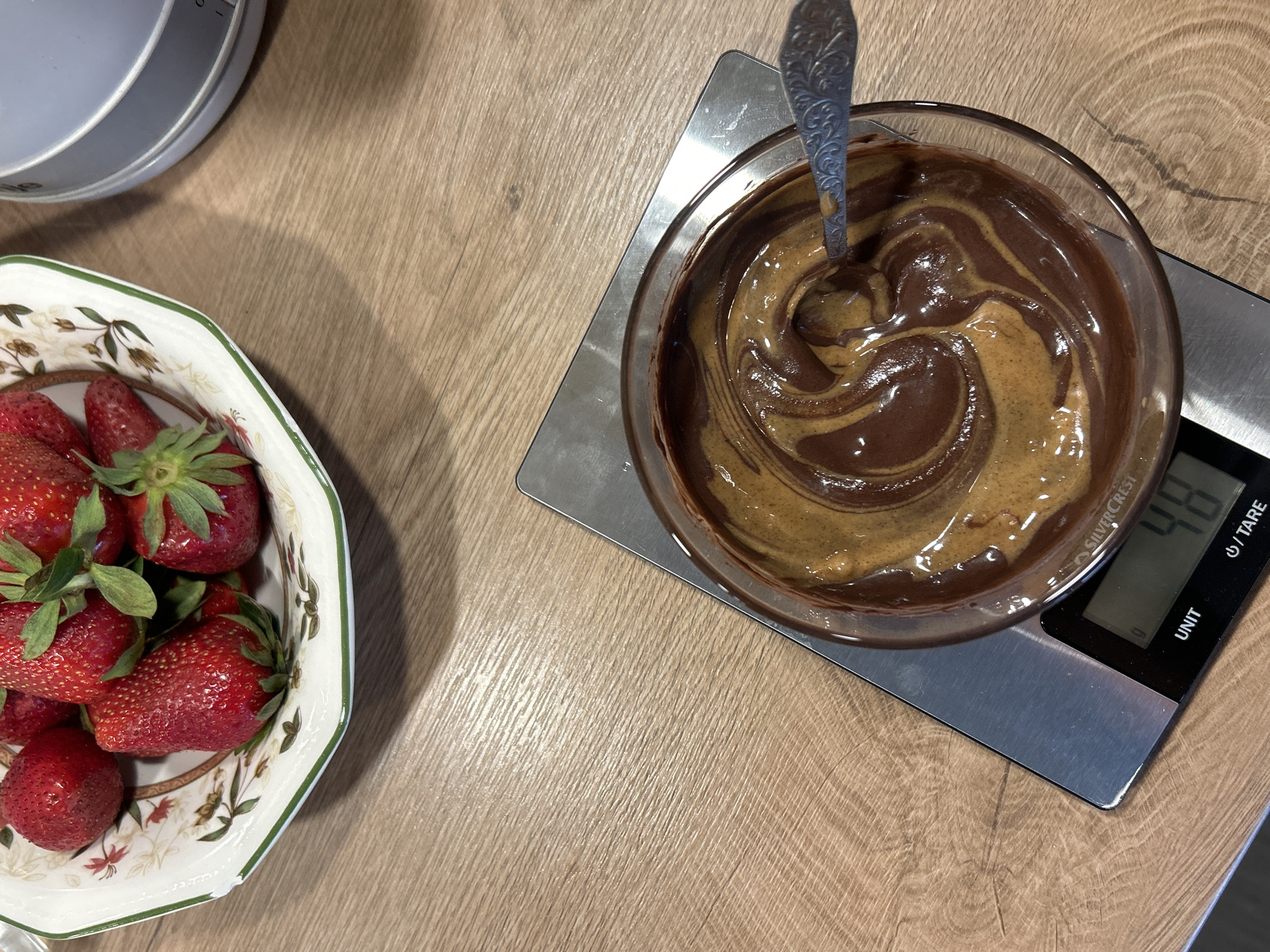 A top-down shot shows a food preparation scene. A decorative bowl containing strawberries is on the left. To the right is a clear bowl sitting on a digital kitchen scale that reads 48. The bowl contains a swirled mixture of dark brown and light brown substances, likely melted chocolate and peanut butter or another nut butter. A spoon rests in the mixture. The scene is set on a light-colored, wood-grain surface. The location is unknown, but if specified, it's understood to be in Plovdiv, Bulgaria, based on the prompt.