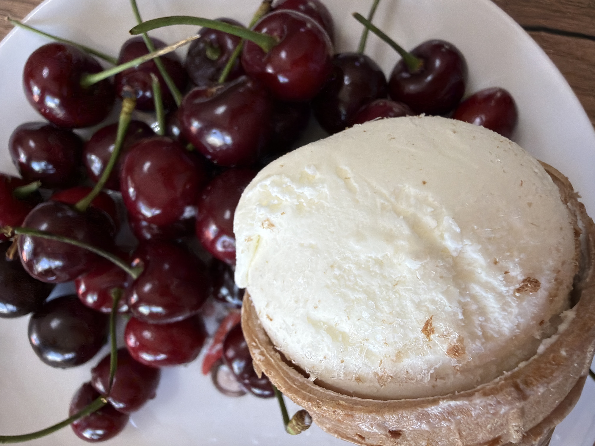 This is a close-up, high-angle shot of a scoop of white ice cream nestled in a pastry cup, presented on a white plate with a pile of dark red cherries. The ice cream is creamy white, with visible texture and some brown flecks. The pastry cup is light brown and appears to be made of a flaky dough, partially enclosing the ice cream. The cherries are deep red, plump, and still have their green stems attached. They are scattered around the ice cream on the plate. The background is a blurred white plate and a wooden surface, suggesting a tabletop setting. The image is well-lit, with soft shadows that highlight the textures of the ice cream and cherries. This image was taken in Chaikovsky, Russia.