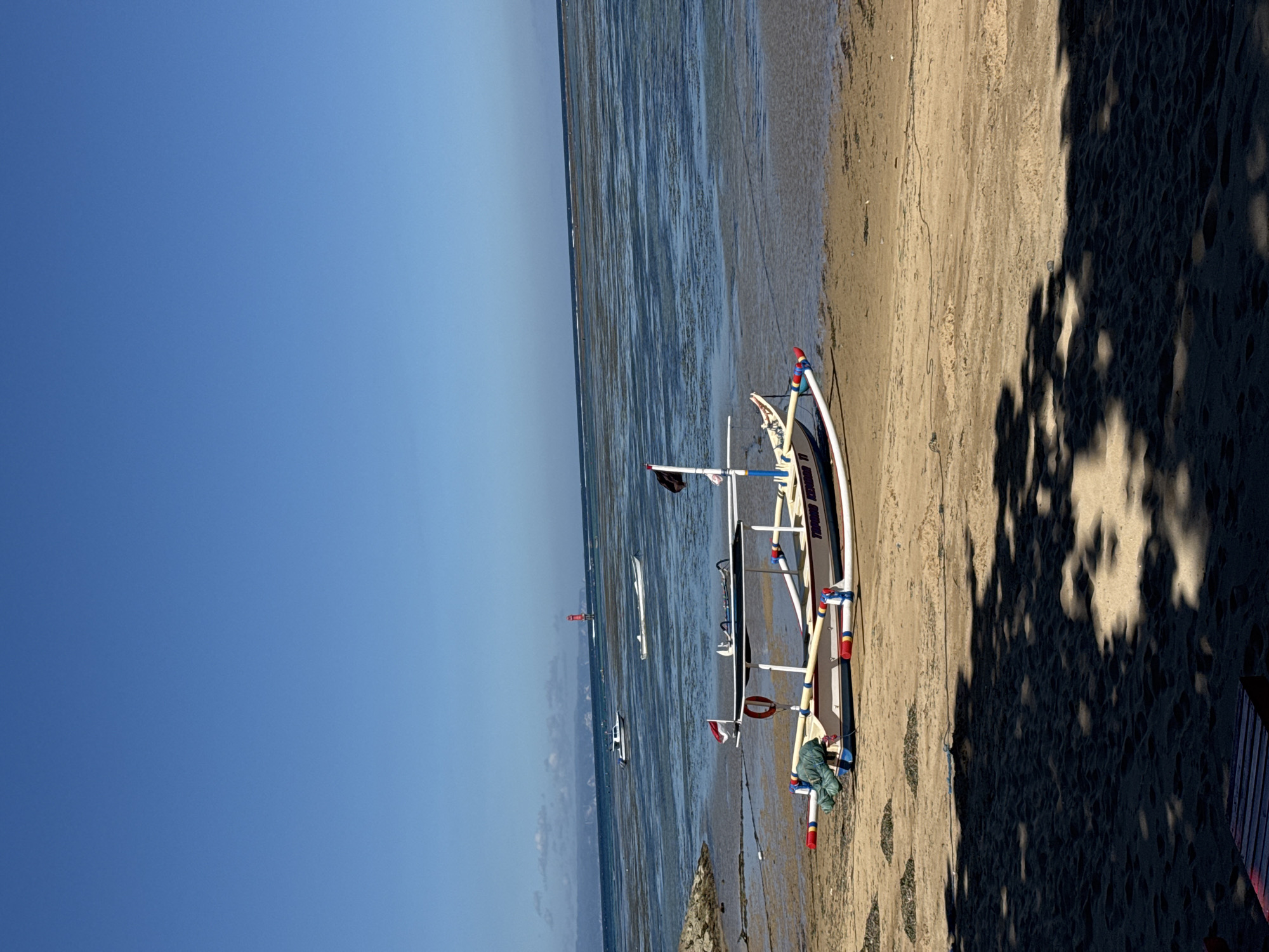 The image captures a serene coastal scene, likely in Denpasar, Indonesia, during daytime. The main subject is a traditional Balinese outrigger fishing boat, known as a Jukung, resting on a sandy beach. The boat is predominantly white with blue accents and red details on its outriggers and structure. On its side, the text "TRUNO JOYO" is clearly visible, with possibly "BALI" or "BAL" also present. A blue cloth or fabric is draped near the stern of the boat.
The Jukung is positioned on the light brown sand, which stretches towards the clear, calm blue ocean. The water appears shallow or at low tide, revealing portions of the seabed with varied textures and colors. In the distance, several other small boats are dotted across the water, including what looks like a red buoy or marker. The sky above is a vibrant, cloudless blue, indicating fair weather.
Strong, dark shadows of unseen trees or foliage fall across the right side of the sandy beach, suggesting the sun is high in the sky, possibly late morning or early afternoon. The overall atmosphere is peaceful and tranquil, typical of a quiet beach setting. There are no people visible in the frame, contributing to the sense of calm.