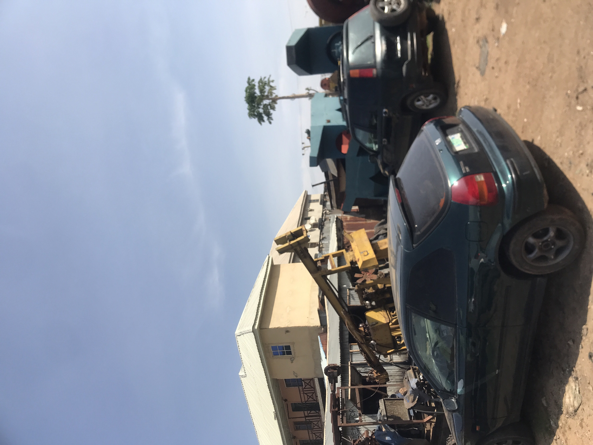 The image depicts an outdoor scene in Rafin Par, Nigeria, likely a commercial or industrial area. In the foreground, a dark green, two-door hatchback car is parked on a dirt surface. Behind it, a dark green SUV is also parked. In the middle ground, a large yellow construction crane or heavy machinery is positioned. Further back, there are several buildings, one of which is a light-colored, multi-story structure with a metal roof. Various other metal structures and what appear to be scrap materials are scattered throughout the scene. A tall, slender tree with green foliage stands in the background to the right of the crane. The sky above is a pale blue with faint clouds. The overall impression is one of a somewhat disorganized outdoor storage or worksite.