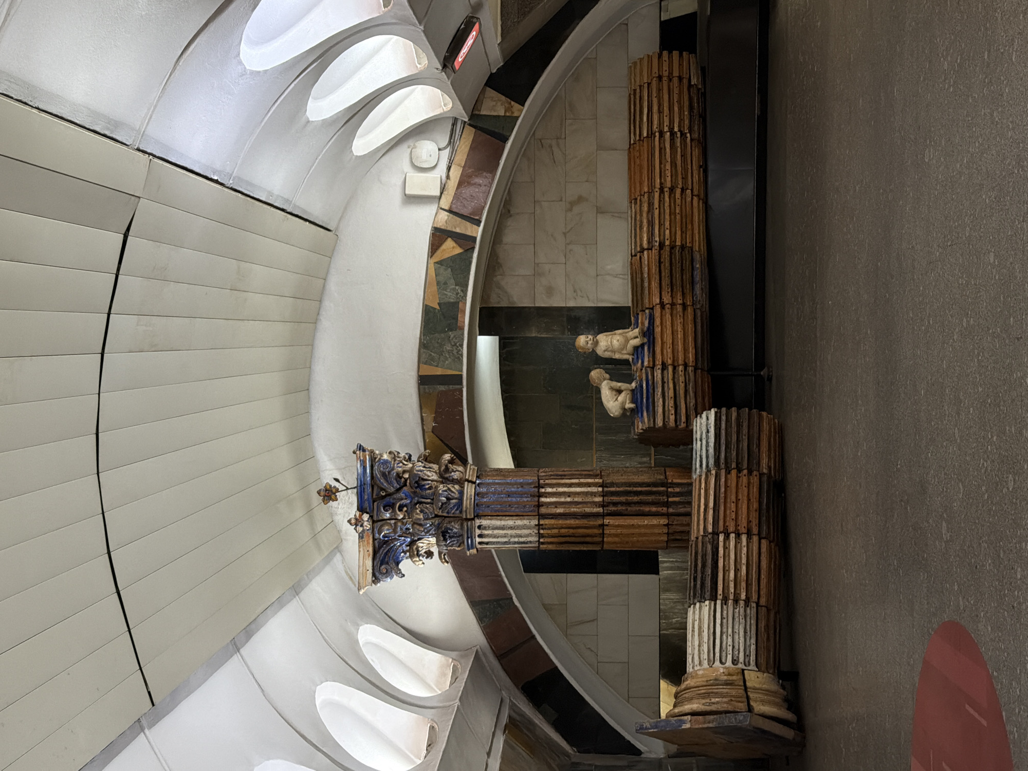 The image depicts a unique architectural detail within the Toledo metro station in Naples, Italy. The station's interior is characterized by a blend of modern and classical elements.
A series of curved white panels form a portion of the ceiling, creating a rhythmic and visually engaging pattern. Adjacent to this ceiling feature, a decorative archway made of multicolored stones adds a layer of texture and historical reference.
The focal point is a pair of stylized, partial columns. Each column is composed of stacked, carved blocks exhibiting alternating colors. Small, sculpted figures of reclining humans are affixed to the upper sections. One column is topped with elaborate, blue-accented ornamentation, while the other has a classical base.