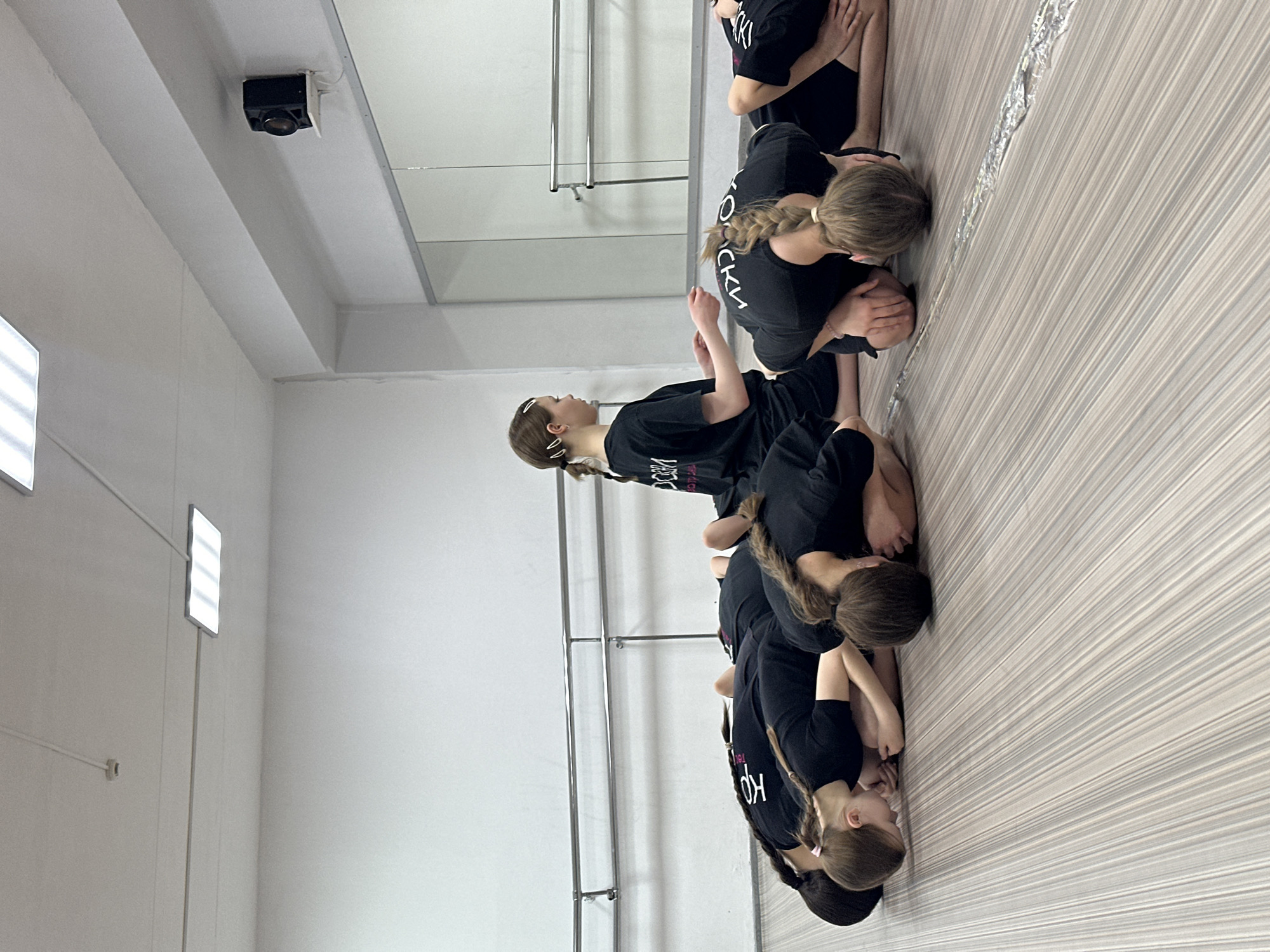 The image displays seven females in what appears to be a dance studio in Votkinsk, Russia. The exact location within Votkinsk is not discernible from visual cues. The studio features white walls, a light-colored floor with thin vertical stripes, a silver-colored double barre mounted along the left wall, and a large mirror occupying the right wall. A black speaker is affixed to the upper right wall, above the mirror. The white ceiling is equipped with rectangular LED light fixtures and a circular smoke detector. All seven individuals are dressed uniformly in black short-sleeved tops, each bearing partially visible Cyrillic text, and black shorts. Six of the females are positioned kneeling on the floor, bent forward with their heads lowered towards the ground, indicating a specific stretch or resting posture. Their hair is primarily styled in braids or ponytails, with one individual wearing white hair clips and another a pink hair clip. The seventh female is seated upright on the floor, facing left, with her hands resting in an open gesture, seemingly observing or instructing the others. A long, narrow strip of crinkled, reflective metallic material is present on the floor in front of the kneeling individuals.