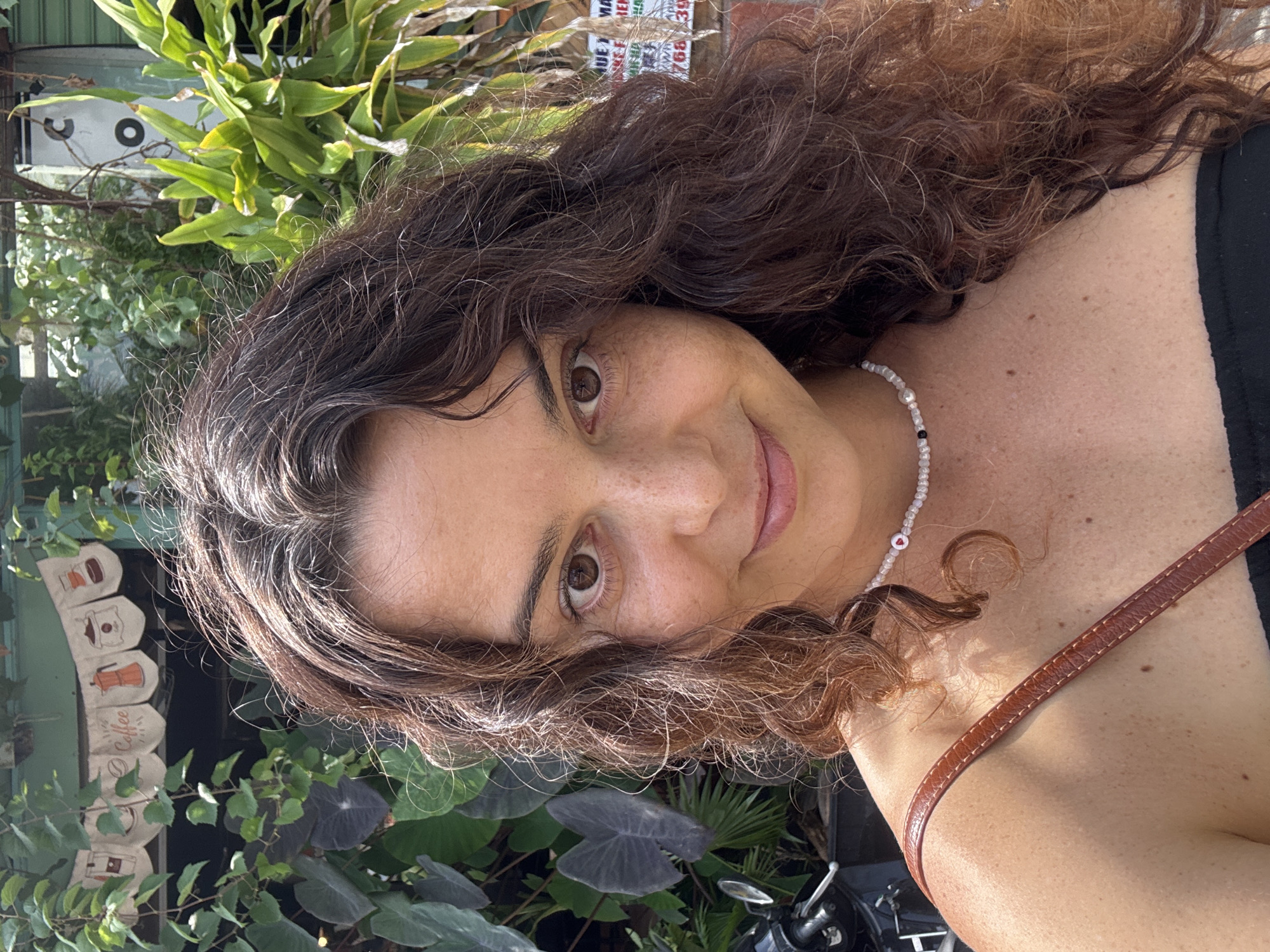 This is a close-up, eye-level, outdoor selfie of a woman with medium-length, wavy, dark brown hair and light brown eyes. She is smiling and has freckles across her nose and cheeks. She is wearing a beaded necklace with a small red heart charm and a black top. A brown strap, possibly from a bag, is visible across her shoulder.
In the background, there are lush green plants and a building with a banner that has illustrations of coffee cups and the word "Coffee" on it. To the right, there's a white sign with some text that is difficult to read, and to the left, the handlebars of a motorcycle are visible. The overall impression is of a casual moment captured in a vibrant, green urban environment, likely in Mũi Né, Vietnam, given the presence of tropical foliage and the style of the signage.