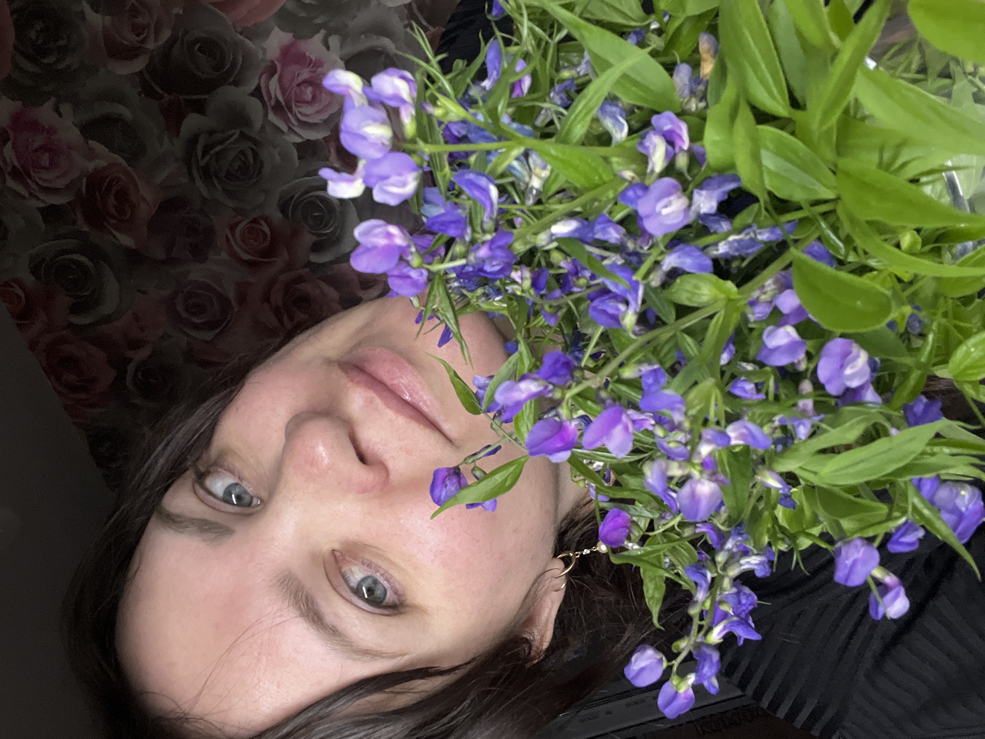 A person with fair skin, brown hair, and light eyes is featured in this close-up shot. They are holding a bunch of purple flowers with green leaves. The background includes a floral pattern with roses in shades of pink and gray. The person is wearing a dark-colored top. The image appears to be taken indoors. There is no indication of the location being in the city of Noviy, Russia.