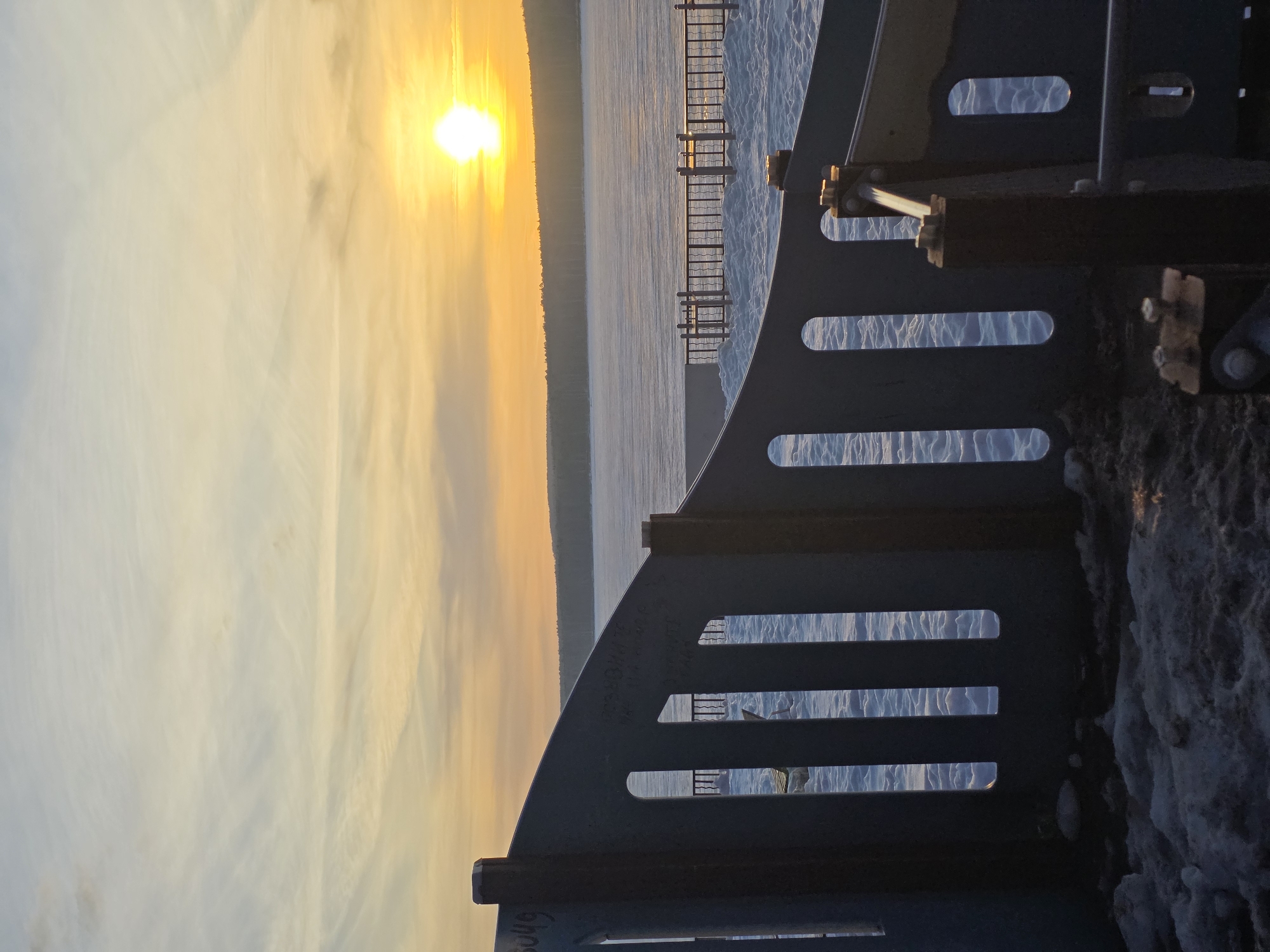 From what I can see, the image appears to be taken from what looks like a portion of the Votkinsk Reservoir, in the city of Chaikovsky, Russia. This is seen through a fence-like structure, the sun is setting over the water in the distance, casting a warm glow on the sky and the reservoir. There are distant landmasses across the water. The immediate foreground shows snow. There is a fence, and some sort of metallic frame.