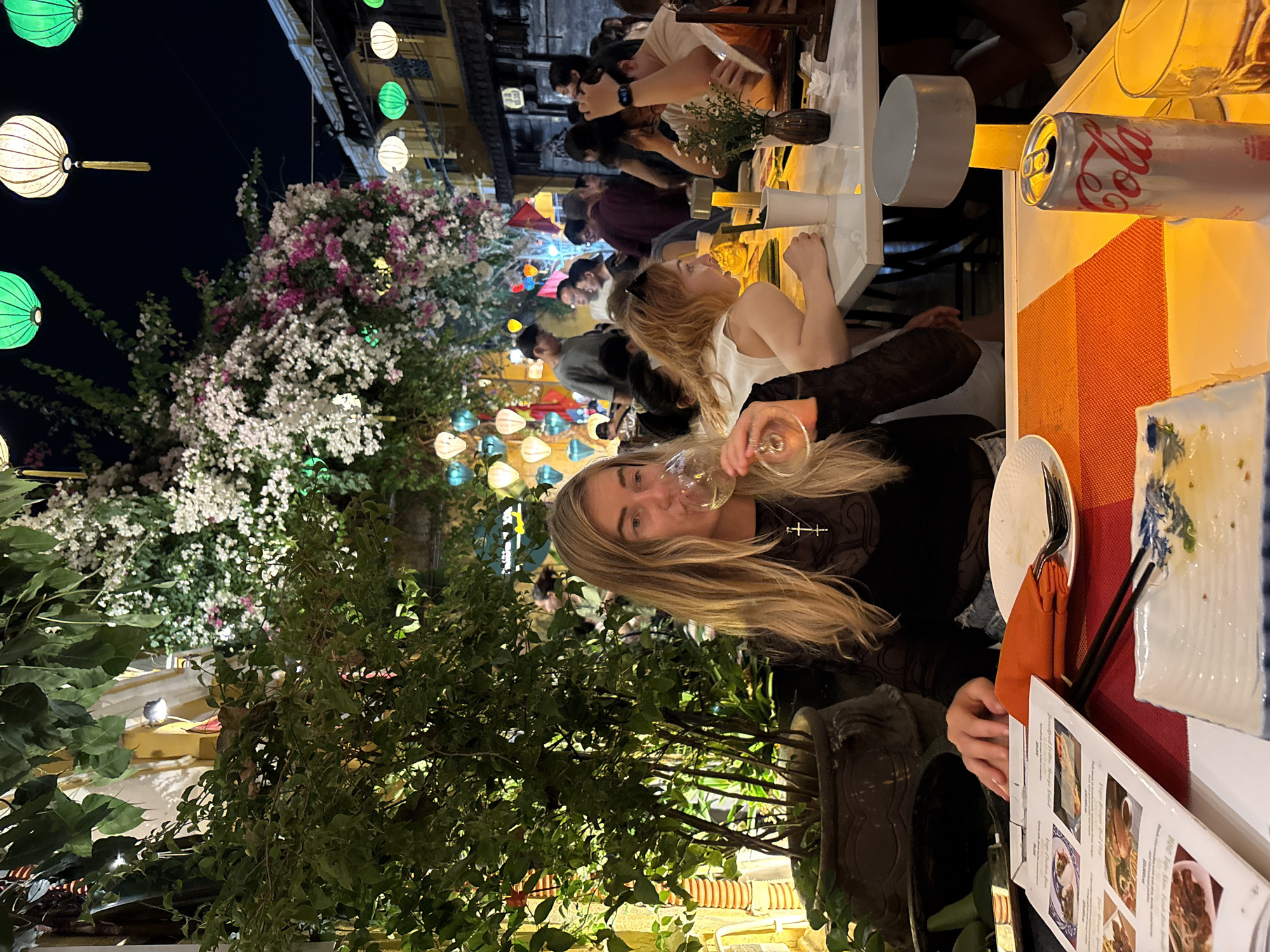 A restaurant scene in Hội An Ward, Vietnam, shows a group of people dining. A woman with blonde hair is drinking from a wine glass. A partially visible Coca-Cola can sits on the table near dishes and cutlery. The background shows lanterns and plants, typical of Hội An's scenery.