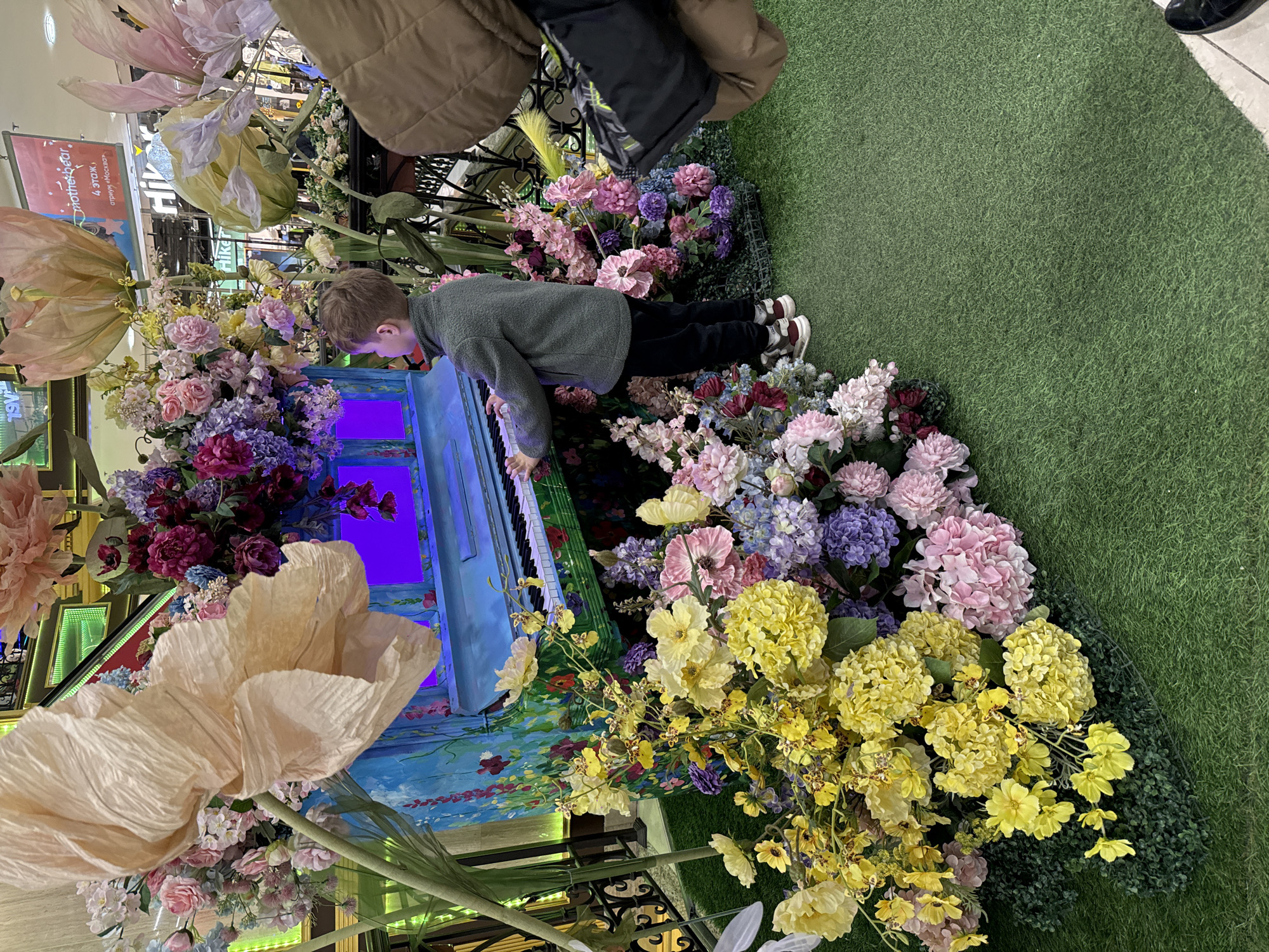 A young boy plays a small, blue piano amidst an elaborate floral display. The piano is decorated with painted flowers, and the area around it is filled with a variety of colorful blossoms, including pink, purple, and yellow flowers. The scene is set on a green, grassy-looking surface, likely artificial turf. Overhead, large, artificial flowers hang, and parts of a building with decorative elements and signs in Russian are visible.
The scene is located in a shopping mall in Moscow, Russia.
